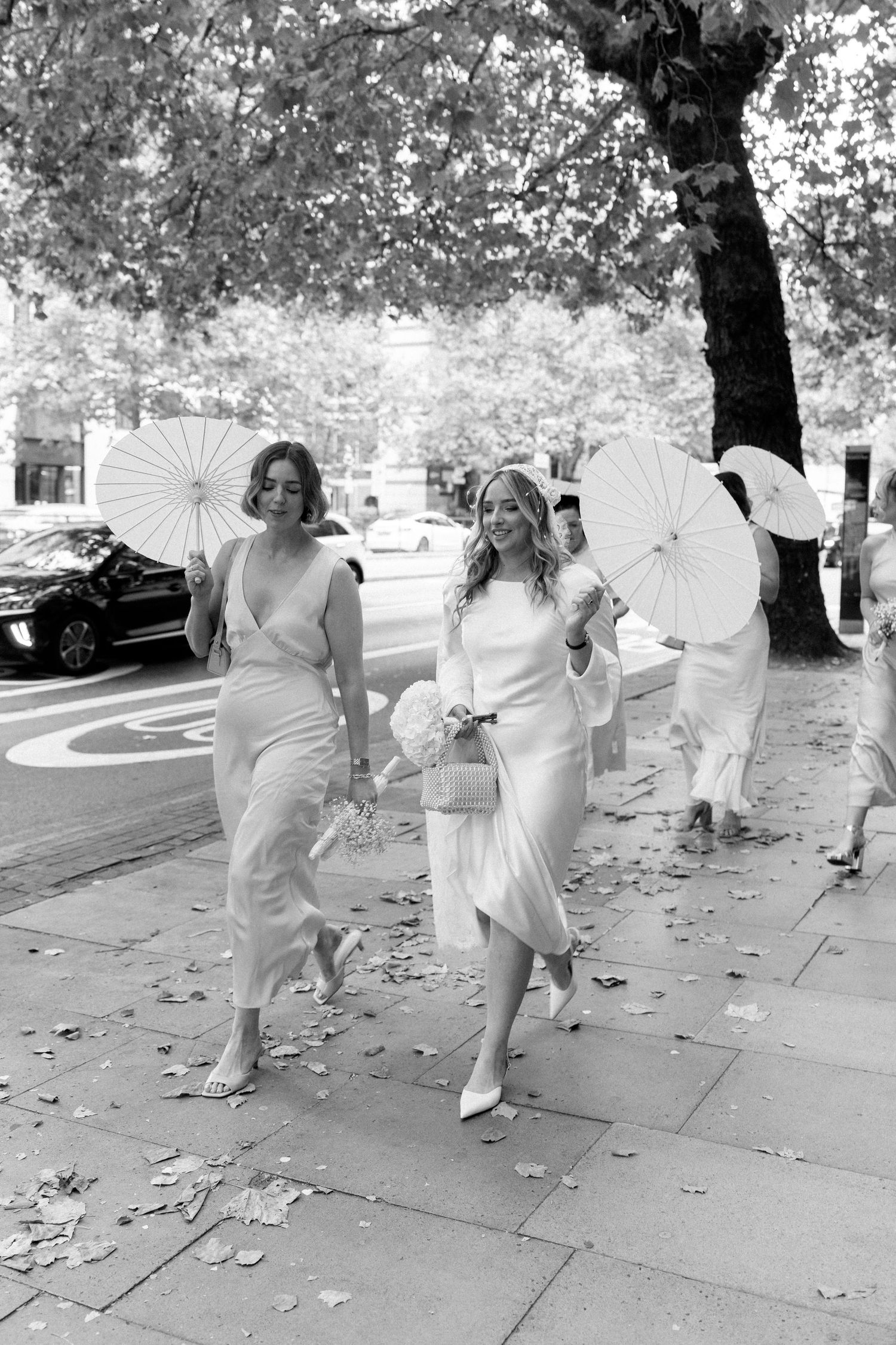 A group of people in white dresses holding white parasols walk together on a tree-lined path.