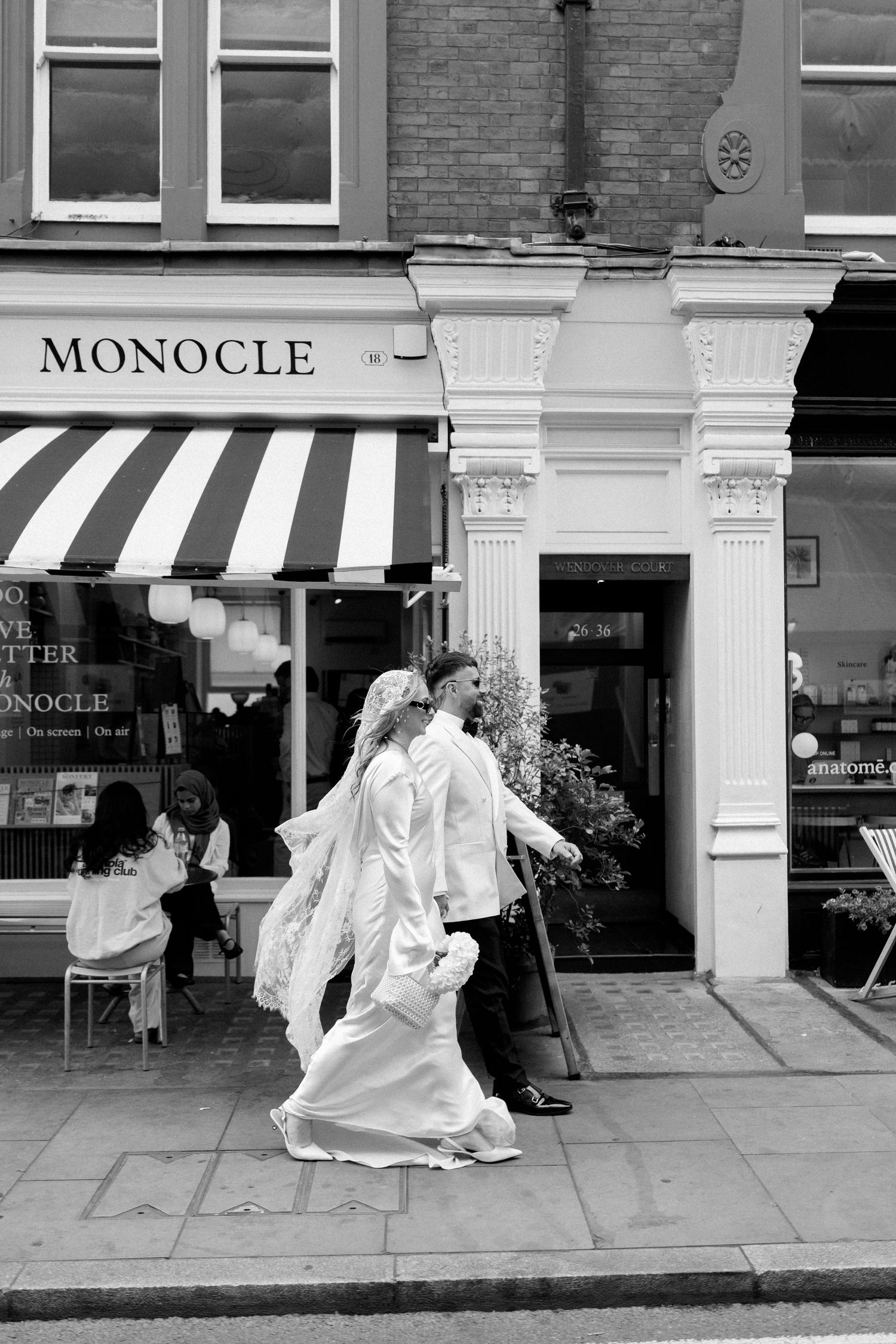 A couple in wedding attire walks past the Monocle store with its distinctive striped awning.