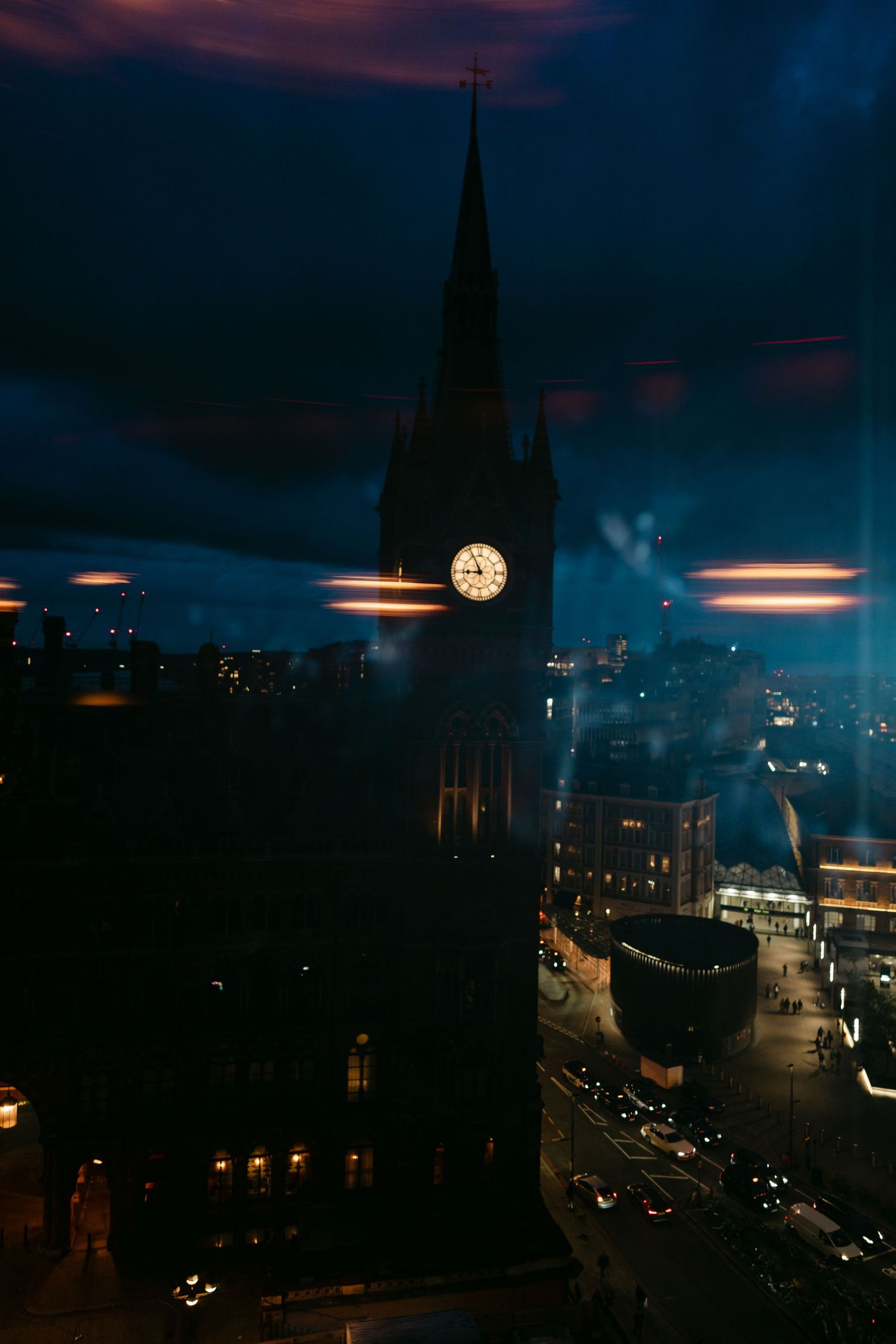 Illuminated church spire with clock tower rises against dark moody sky over city streets at night.