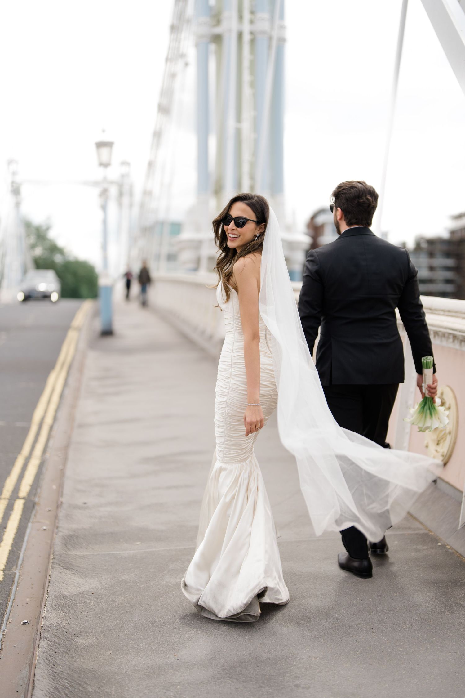 A couple in wedding attire walks along a path with the bride's dress flowing behind them.