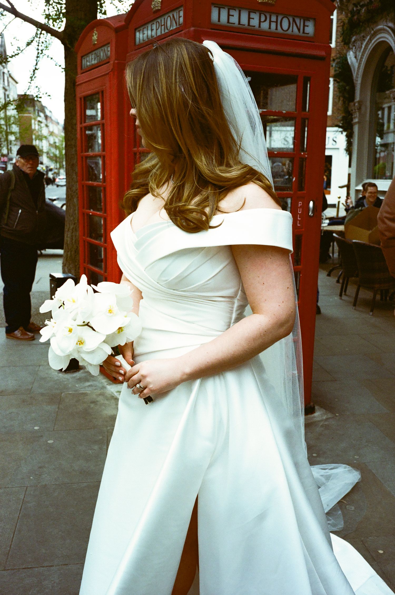 A bride in a white dress stands near iconic red British telephone booths on a London street.