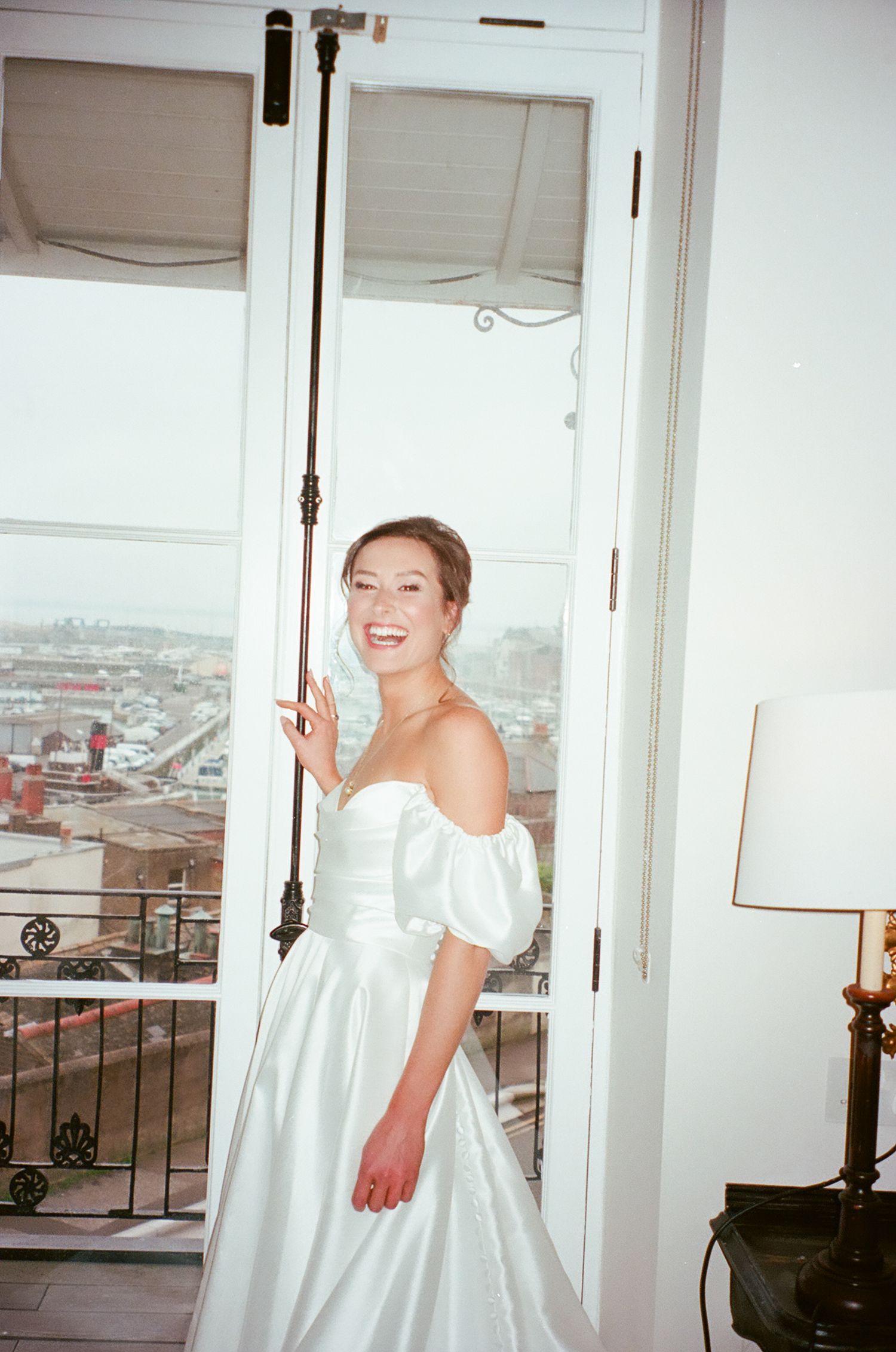 A person in a white off-shoulder wedding dress stands by a window overlooking a city view.