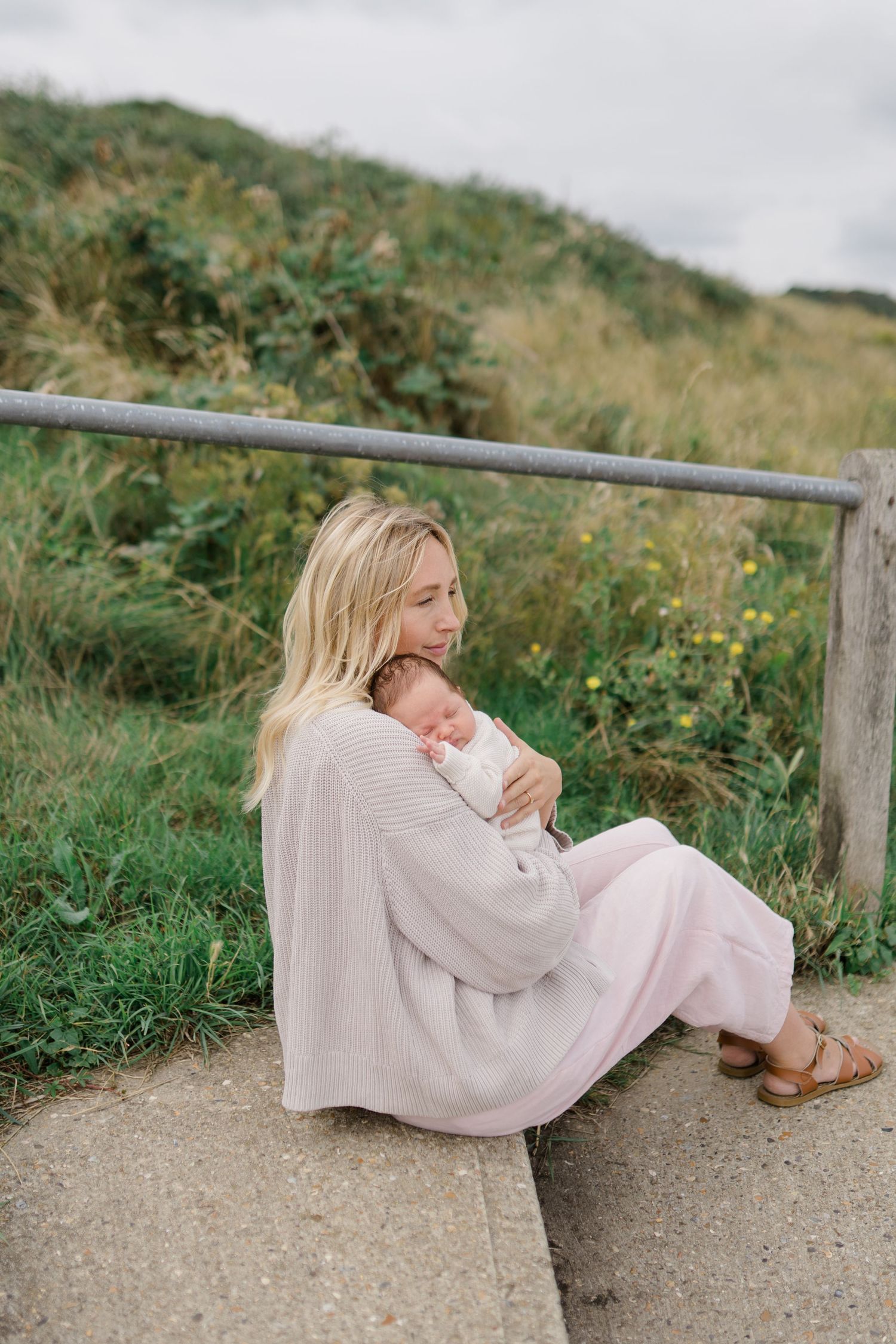 Parent and child rest together on outdoor path surrounded by green coastal vegetation.
