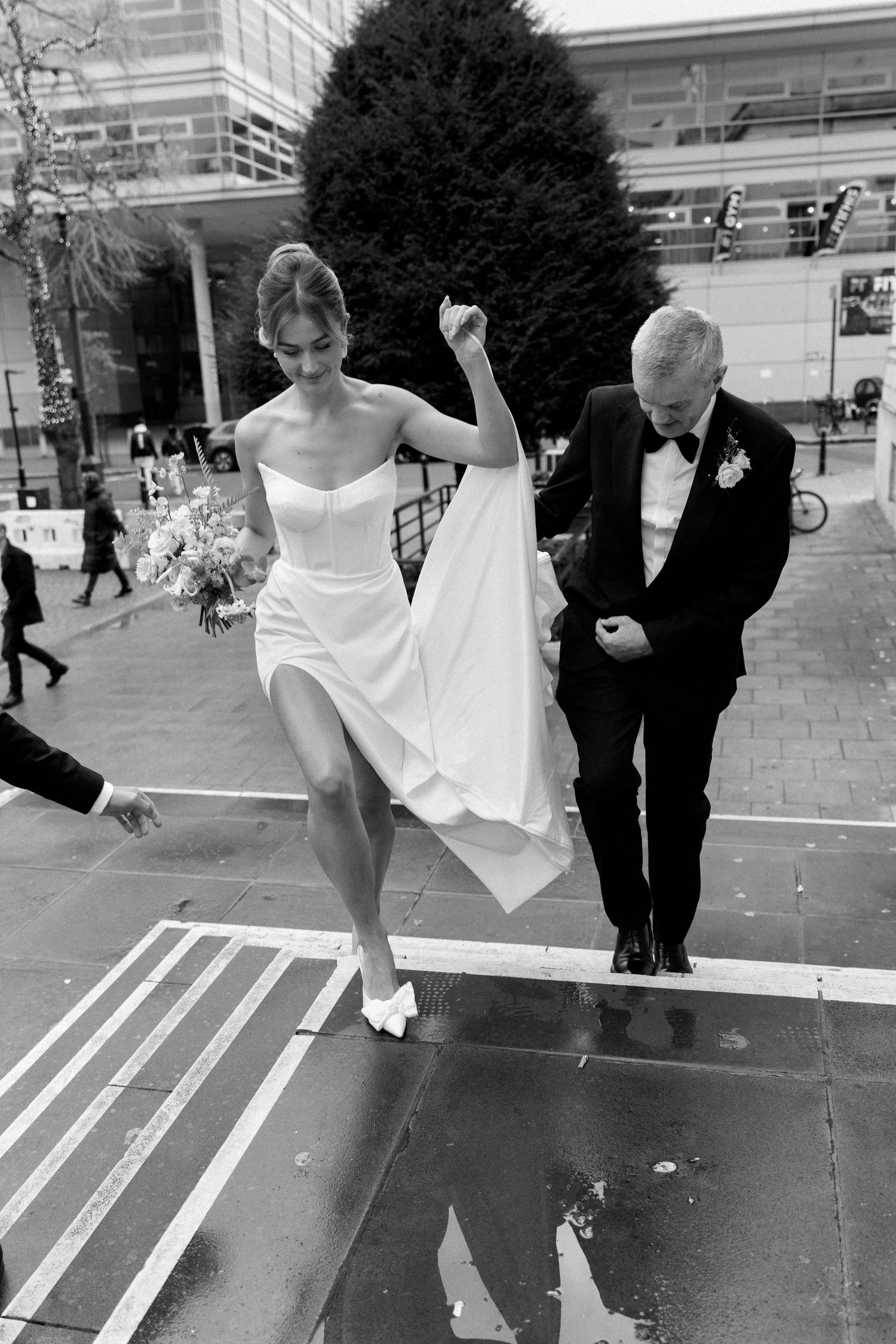 Black and white photo of a couple in wedding attire stepping up onto a platform together outdoors.