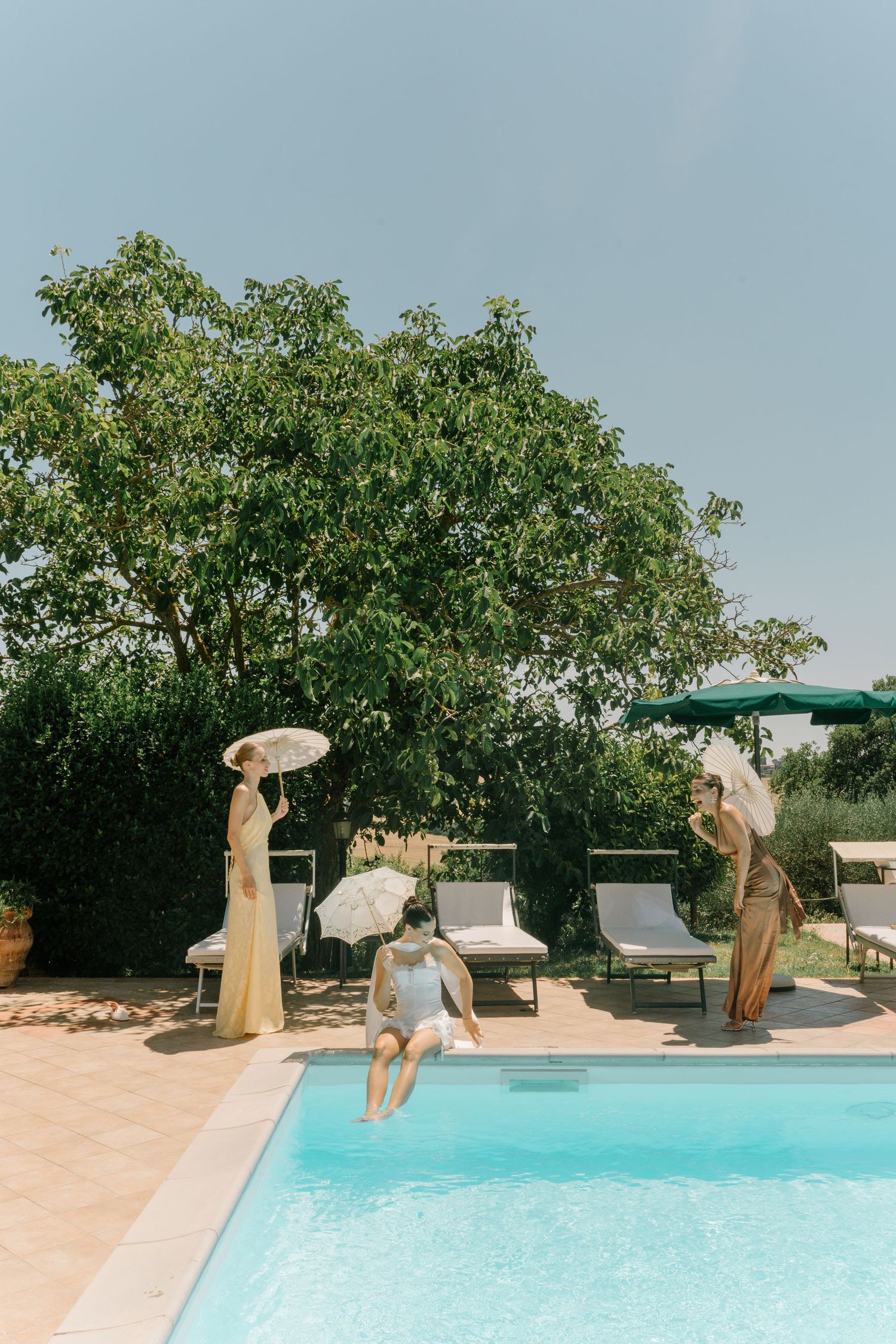 People with parasols enjoying a summer day by a turquoise swimming pool surrounded by lounge chairs and lush trees.