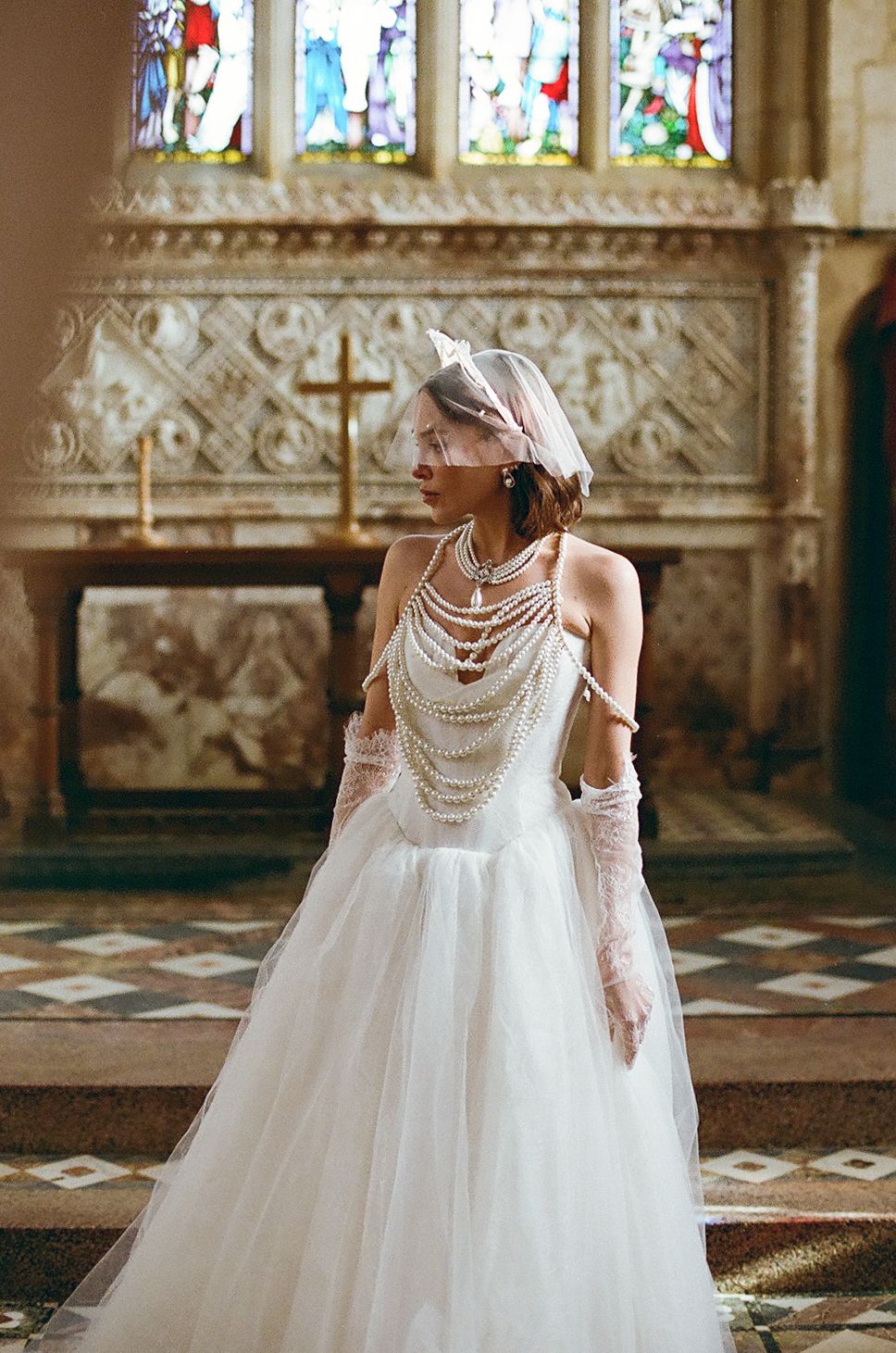 A bride in a flowing white tulle gown with pearl neckline details poses in an ornate church interior.