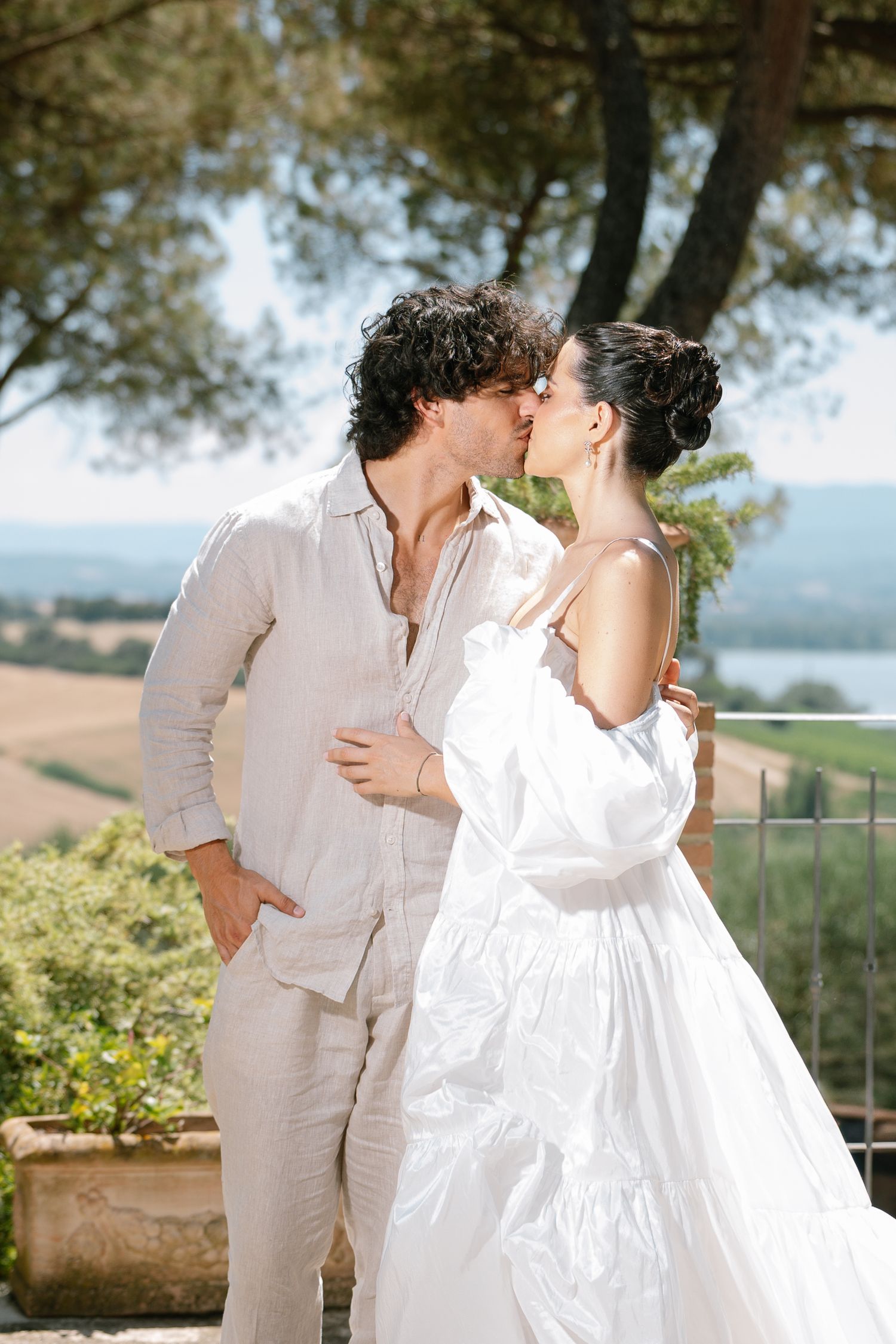 A couple in all-white outfits share an intimate moment against a scenic countryside backdrop.