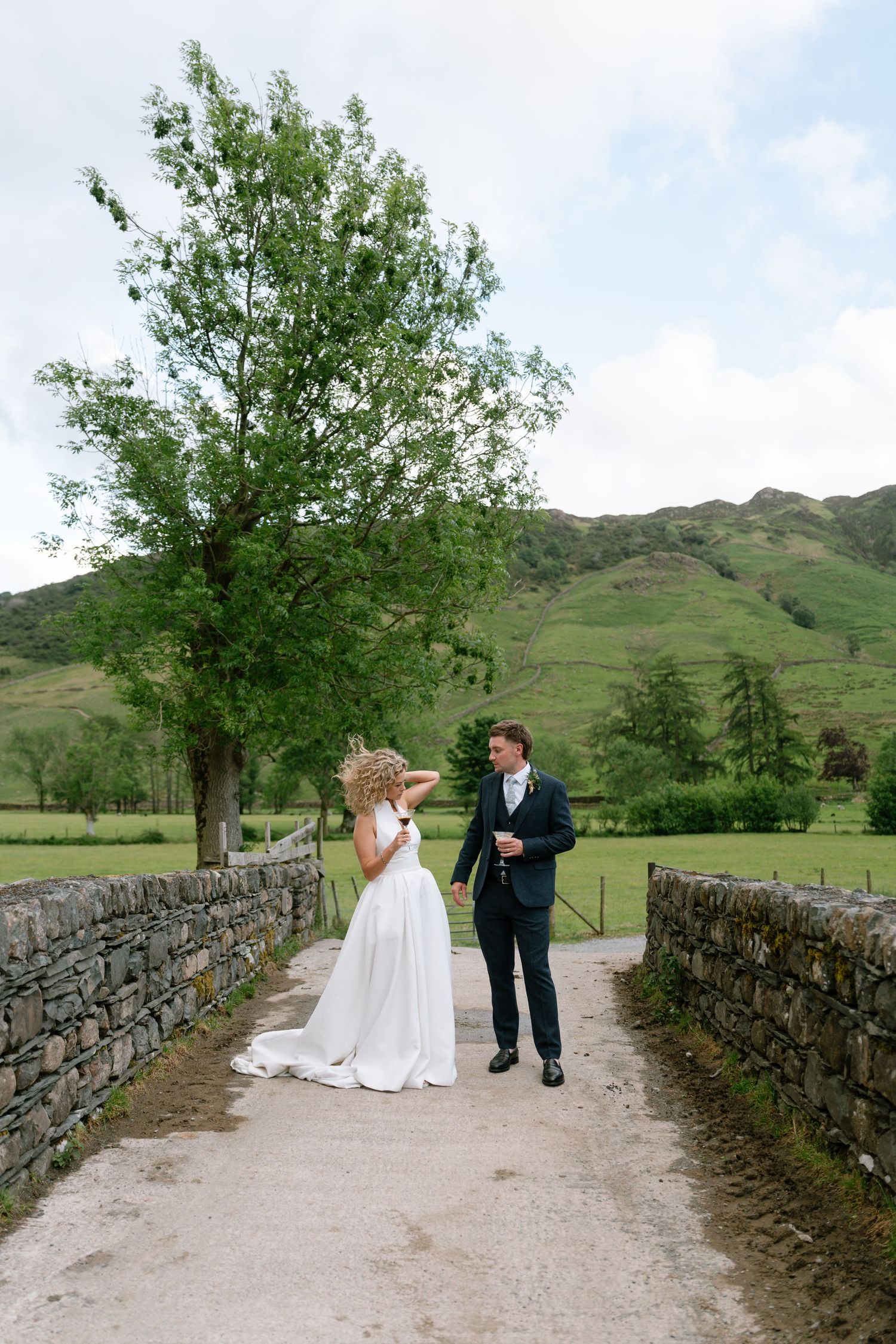 Wedding couple shares a moment on a stone-walled pathway surrounded by green hills.
