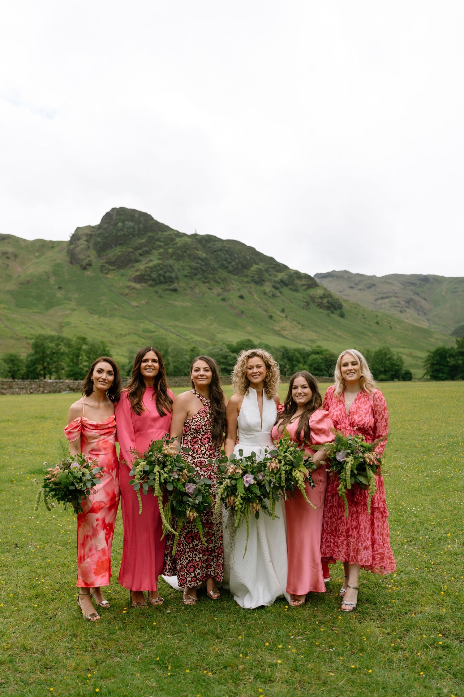 Bridesmaids in coral and floral dresses pose together on green hillside.