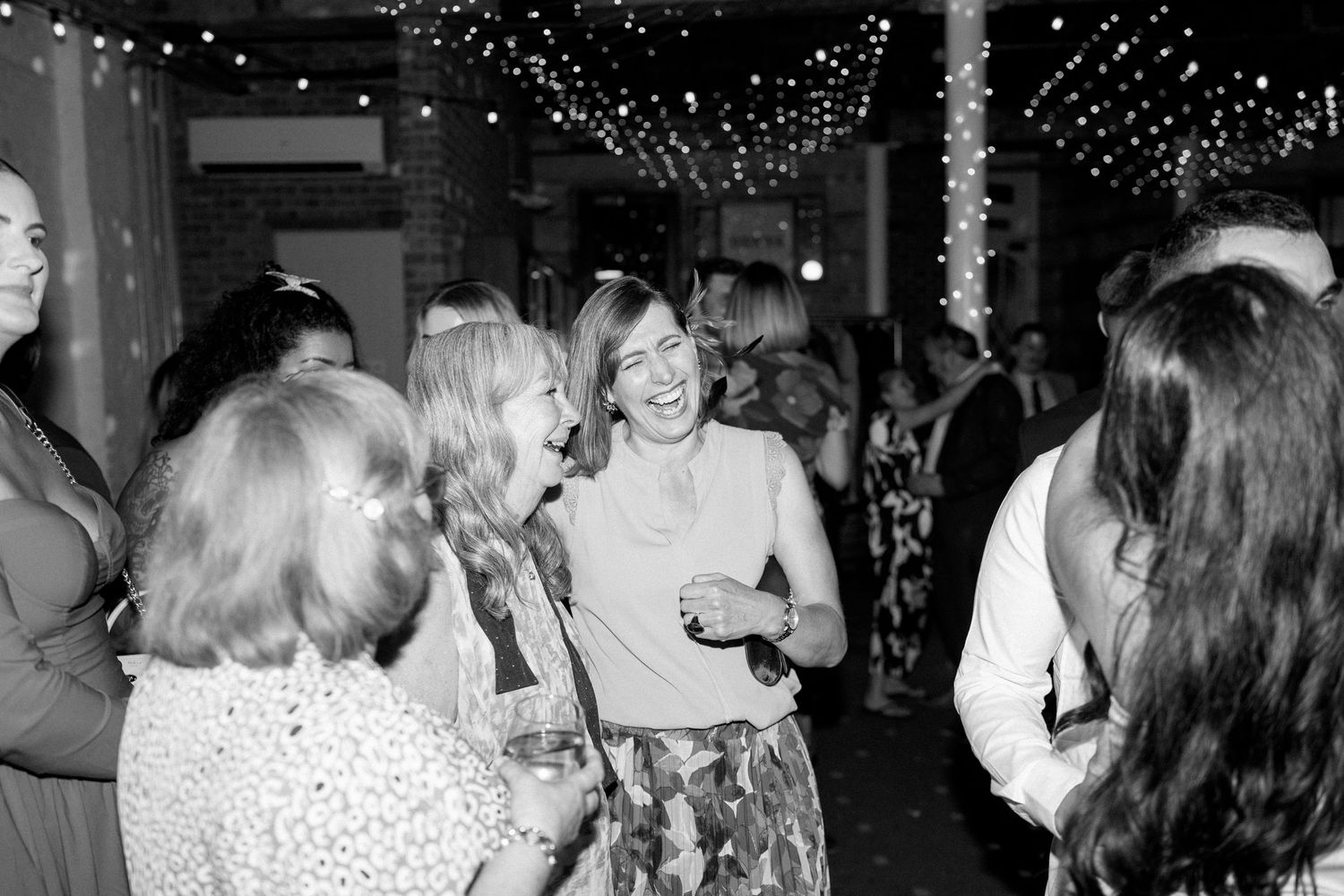 Wedding guests socializing under twinkling lights at an indoor reception.