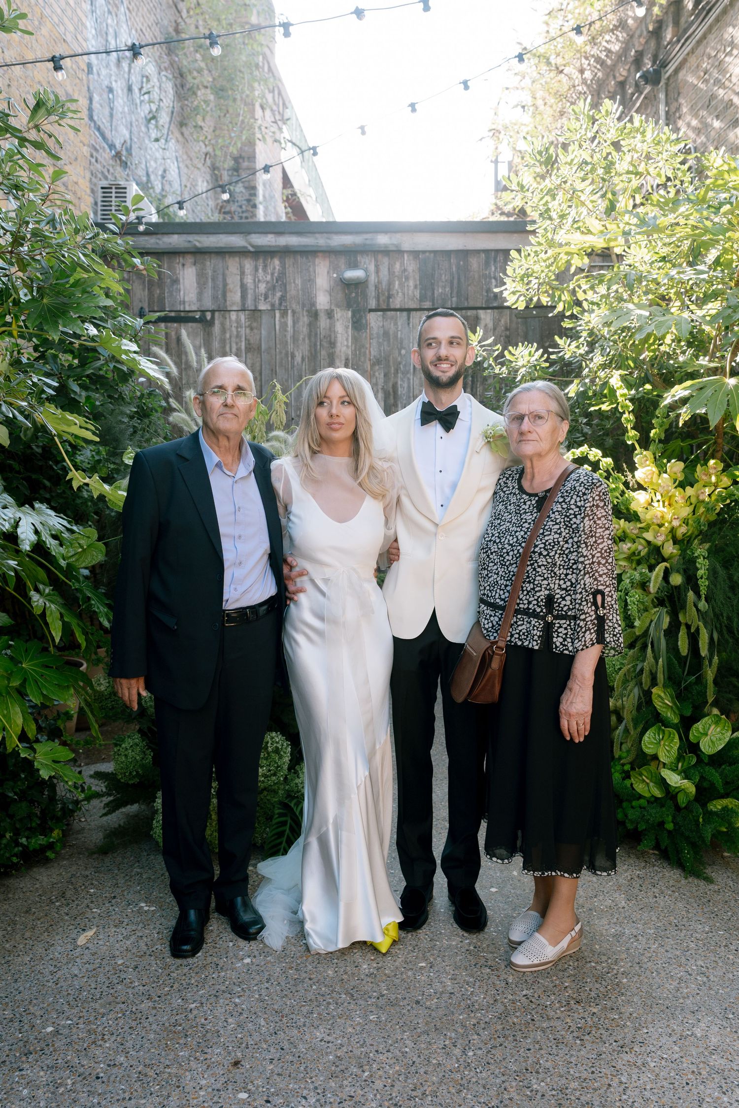 Wedding party poses outdoors in formal attire with greenery backdrop.