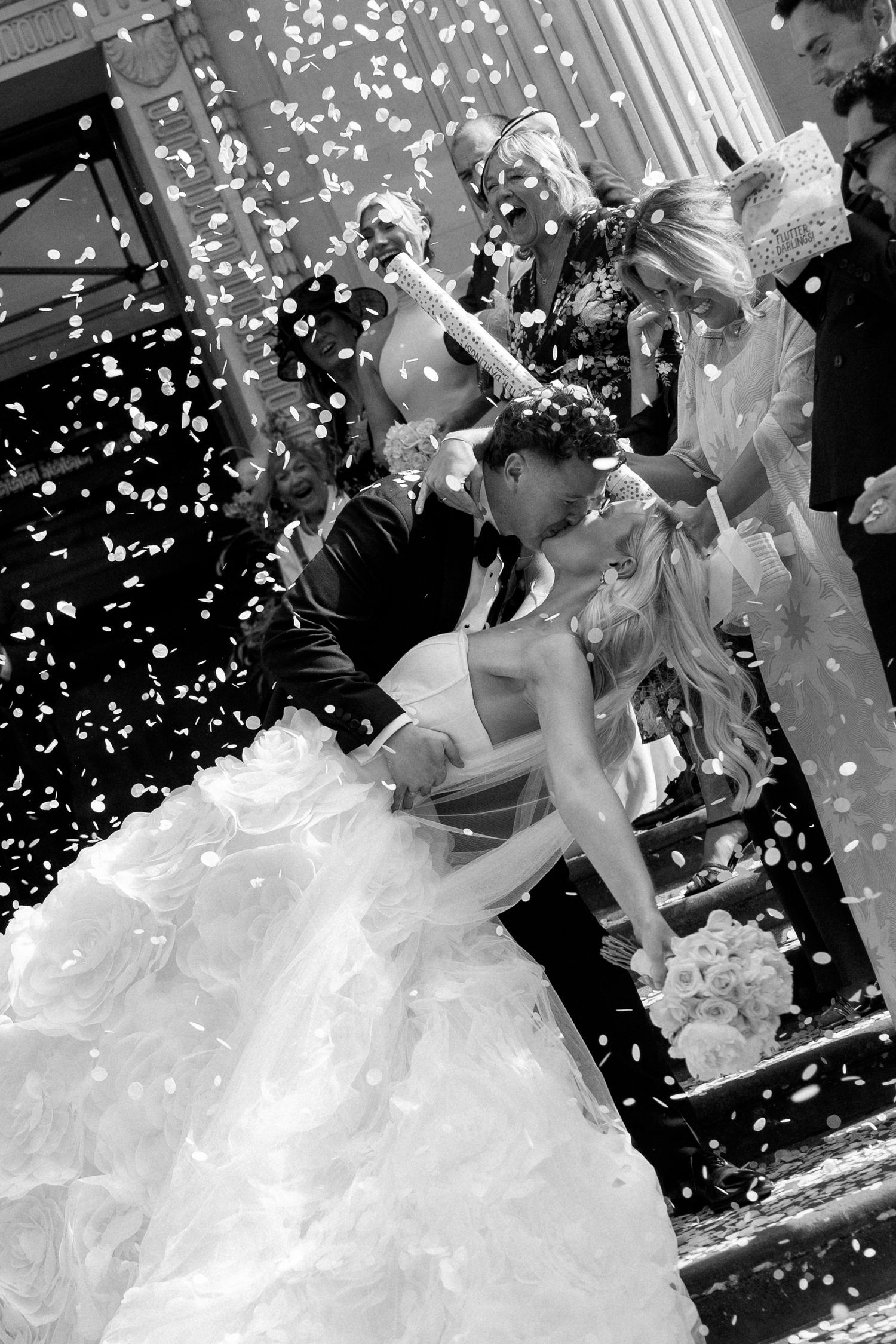 Black and white photo of a bride and groom kissing as confetti falls around them outside a church.