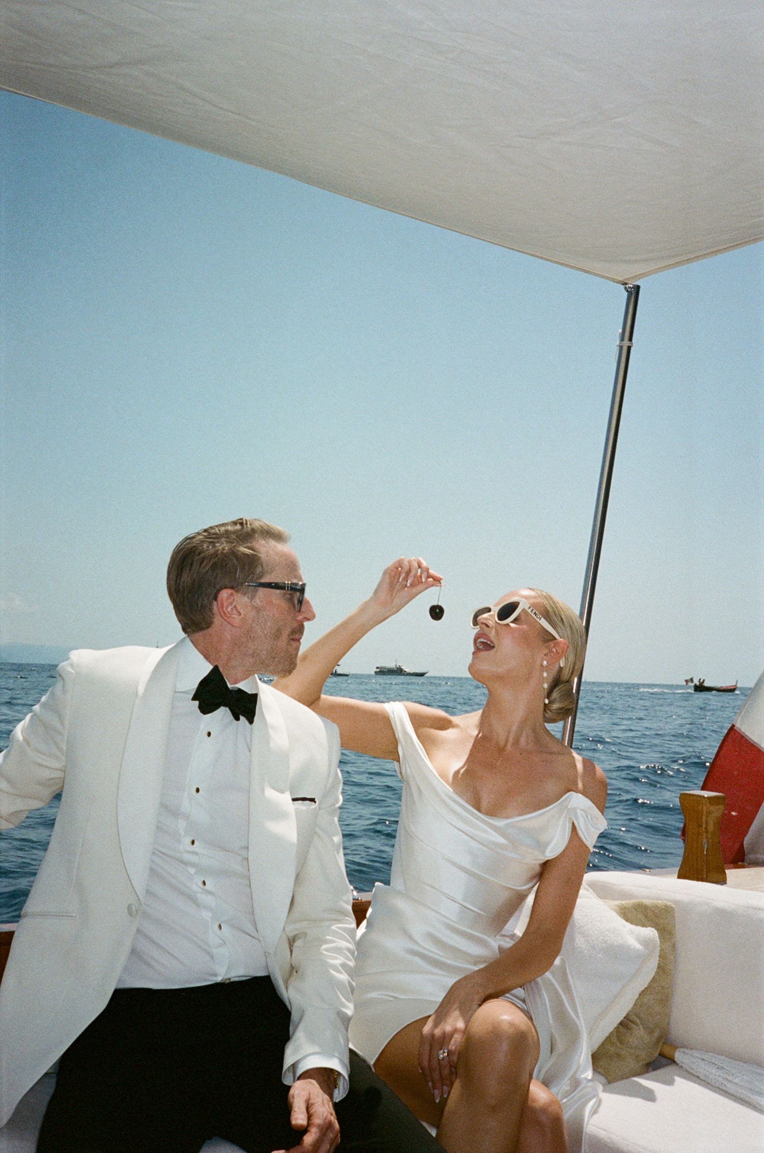 Couple in white wedding attire on a luxury boat, man in tuxedo feeding woman a grape over blue ocean.