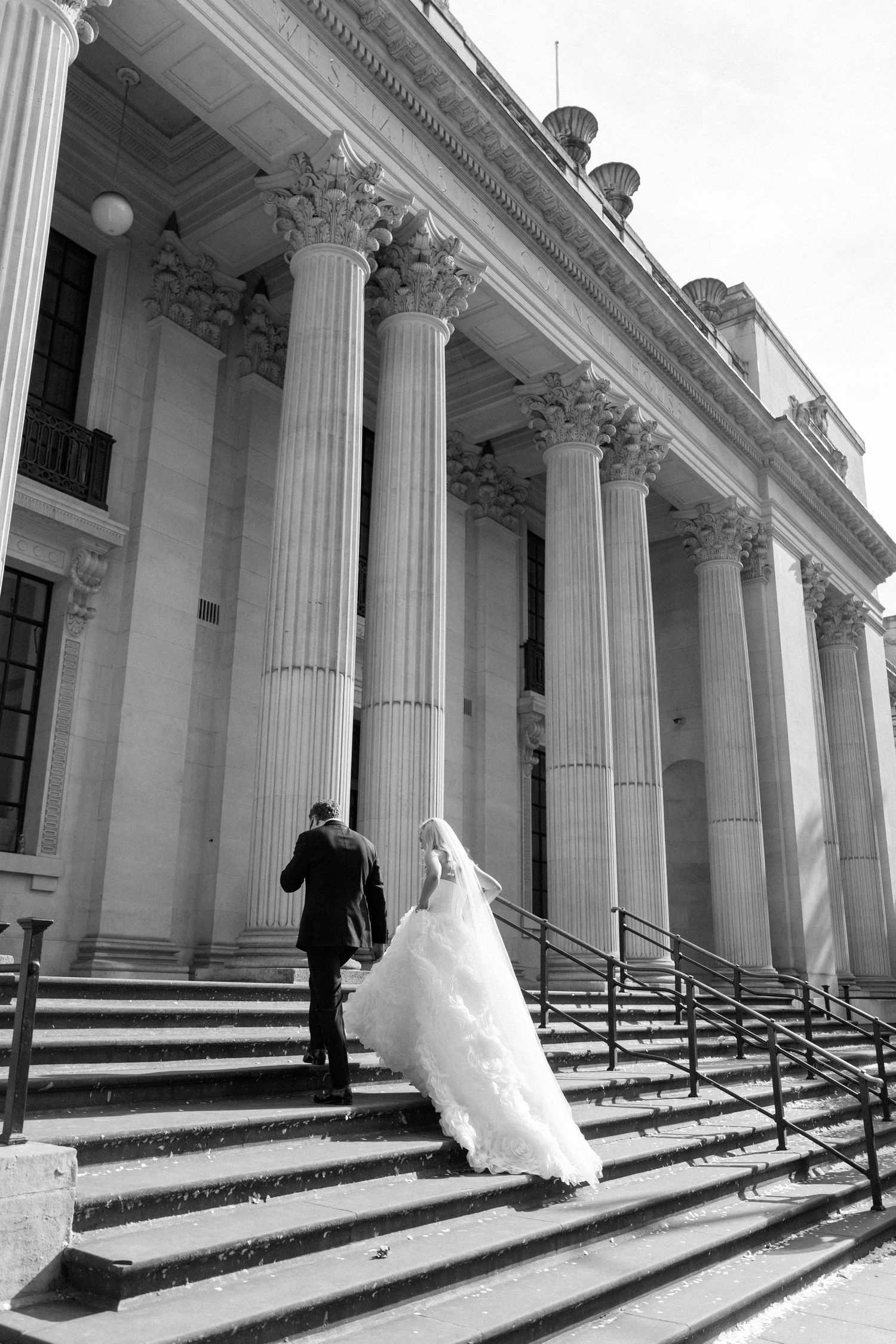 Black and white photo of a bride and groom ascending grand stone steps of a classical building with tall columns.
