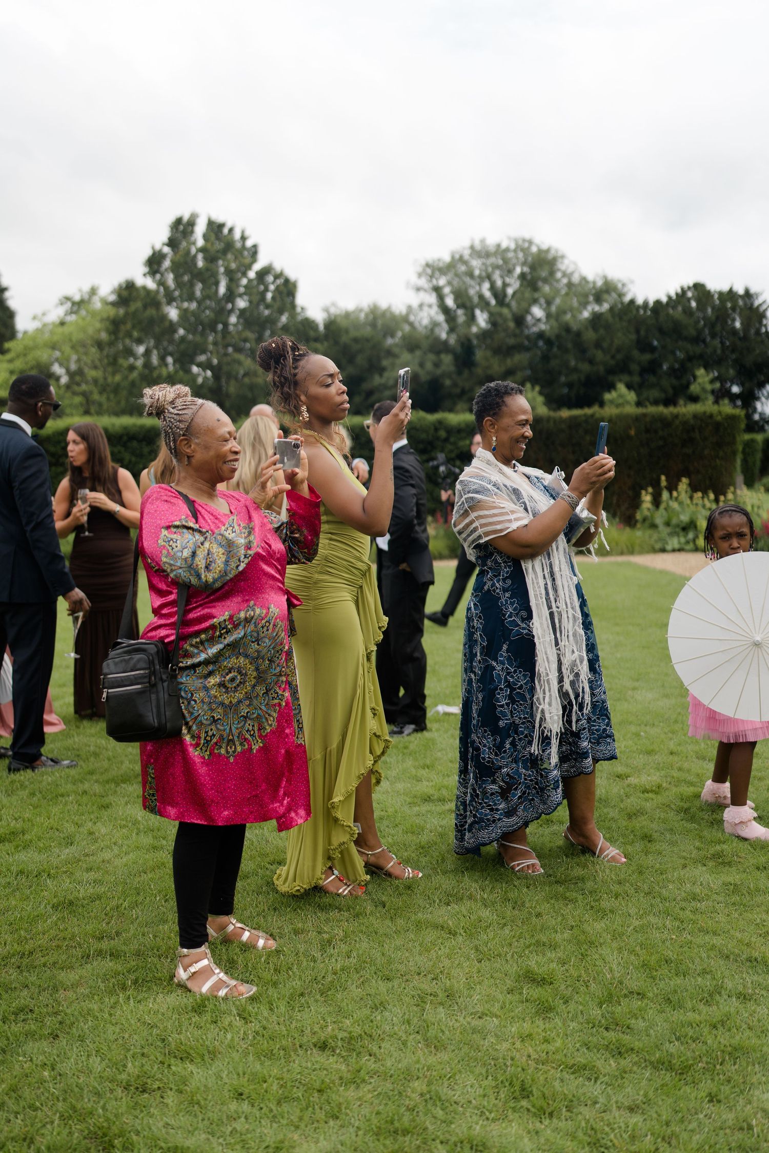 Three women in colorful outfits take photos on their phones at an outdoor wedding reception.
