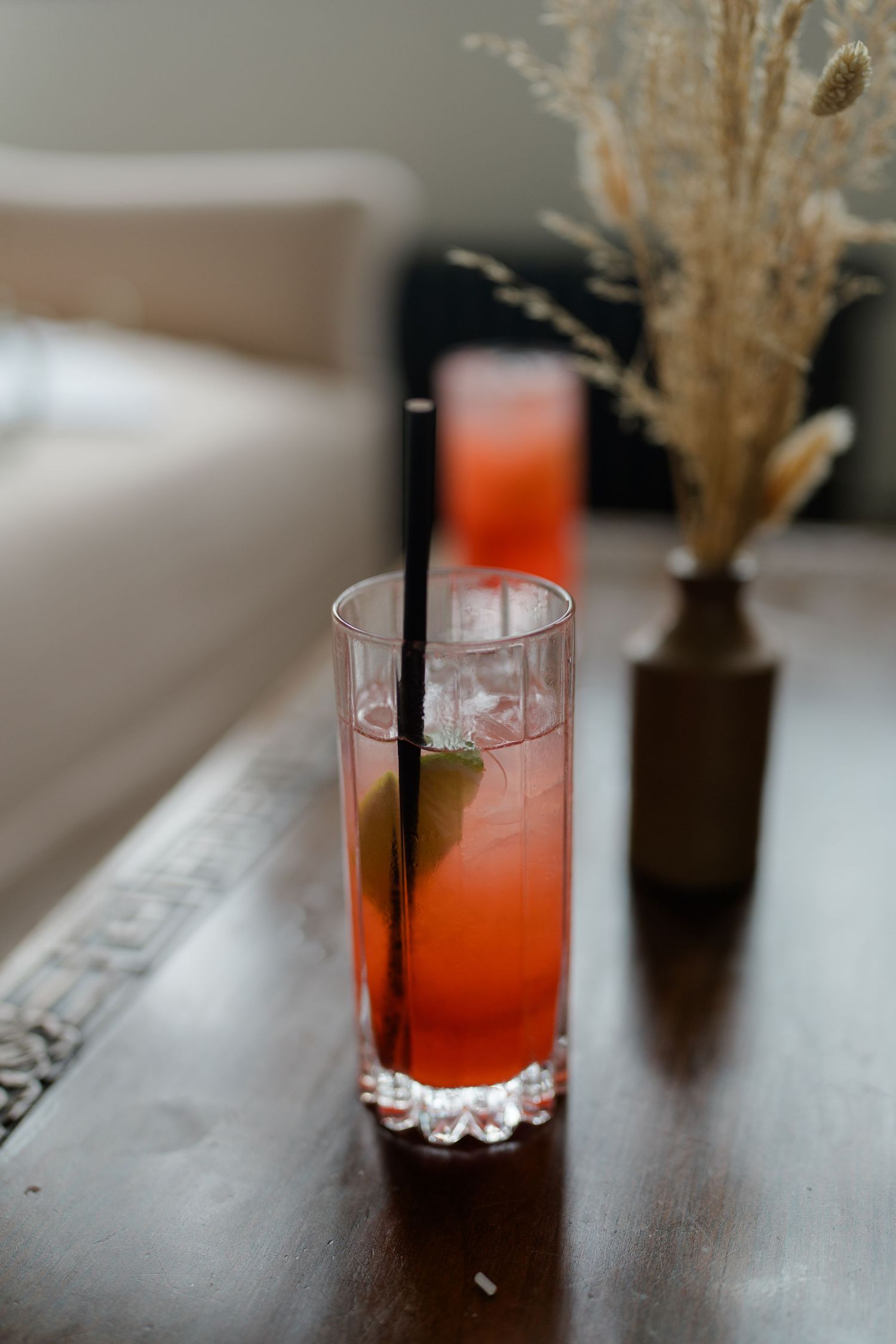 A vibrant red cocktail with ice, lime, and a black straw in a tall glass on a wooden table.