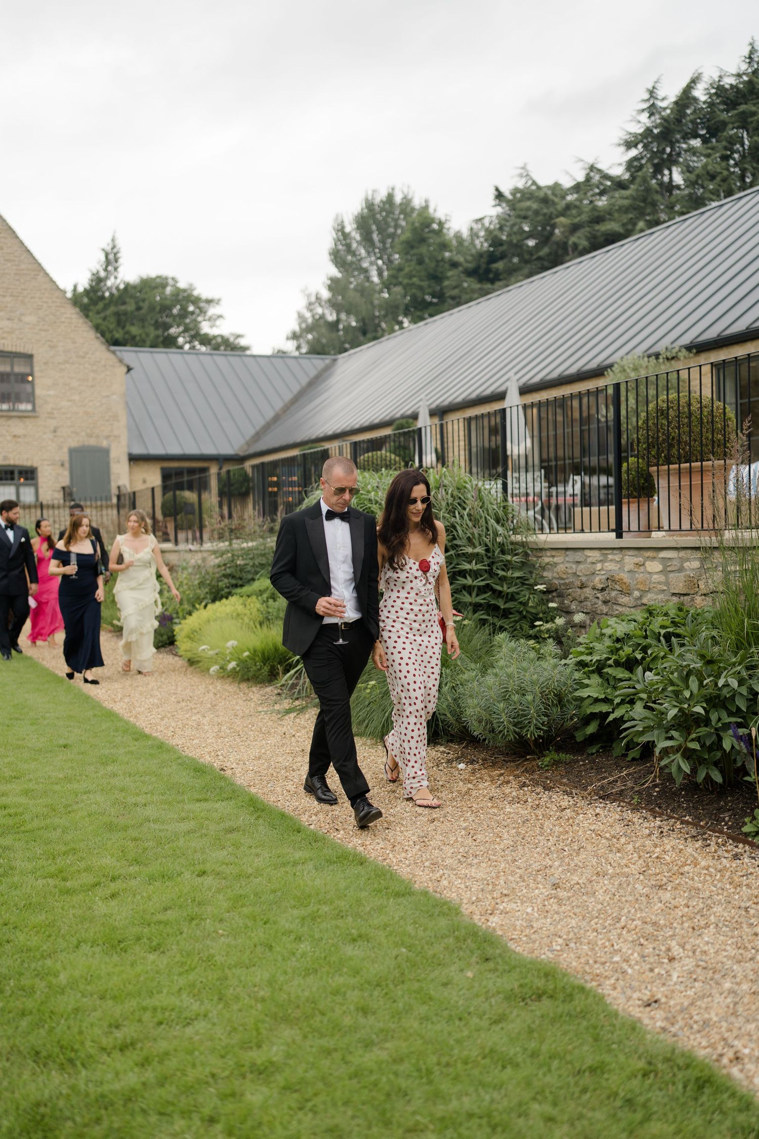 Elegant wedding guests in formal attire walking along a gravel path beside lush gardens at a countryside venue.
