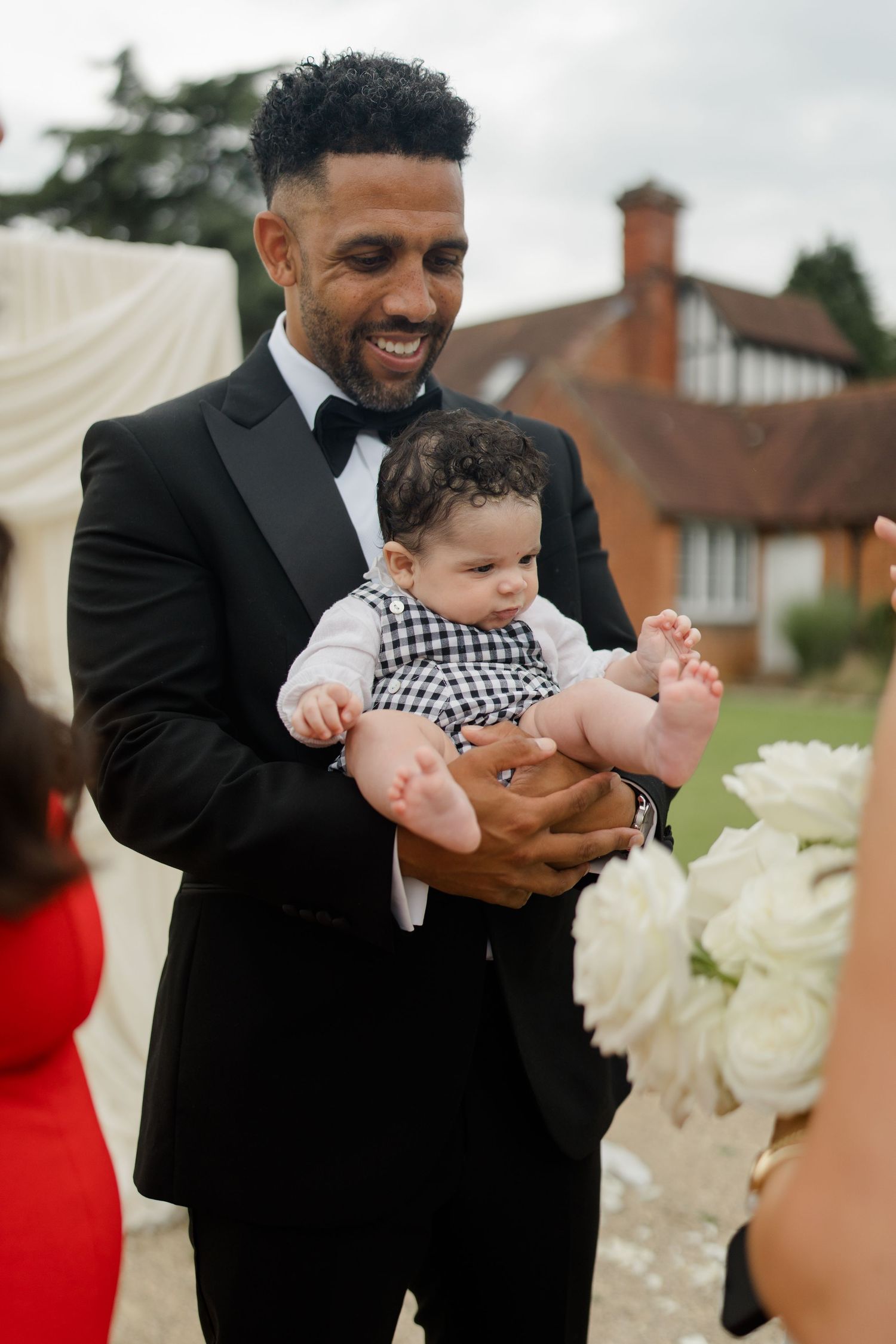 Smiling groom in black tuxedo holding adorable baby in checkered outfit at outdoor wedding reception.