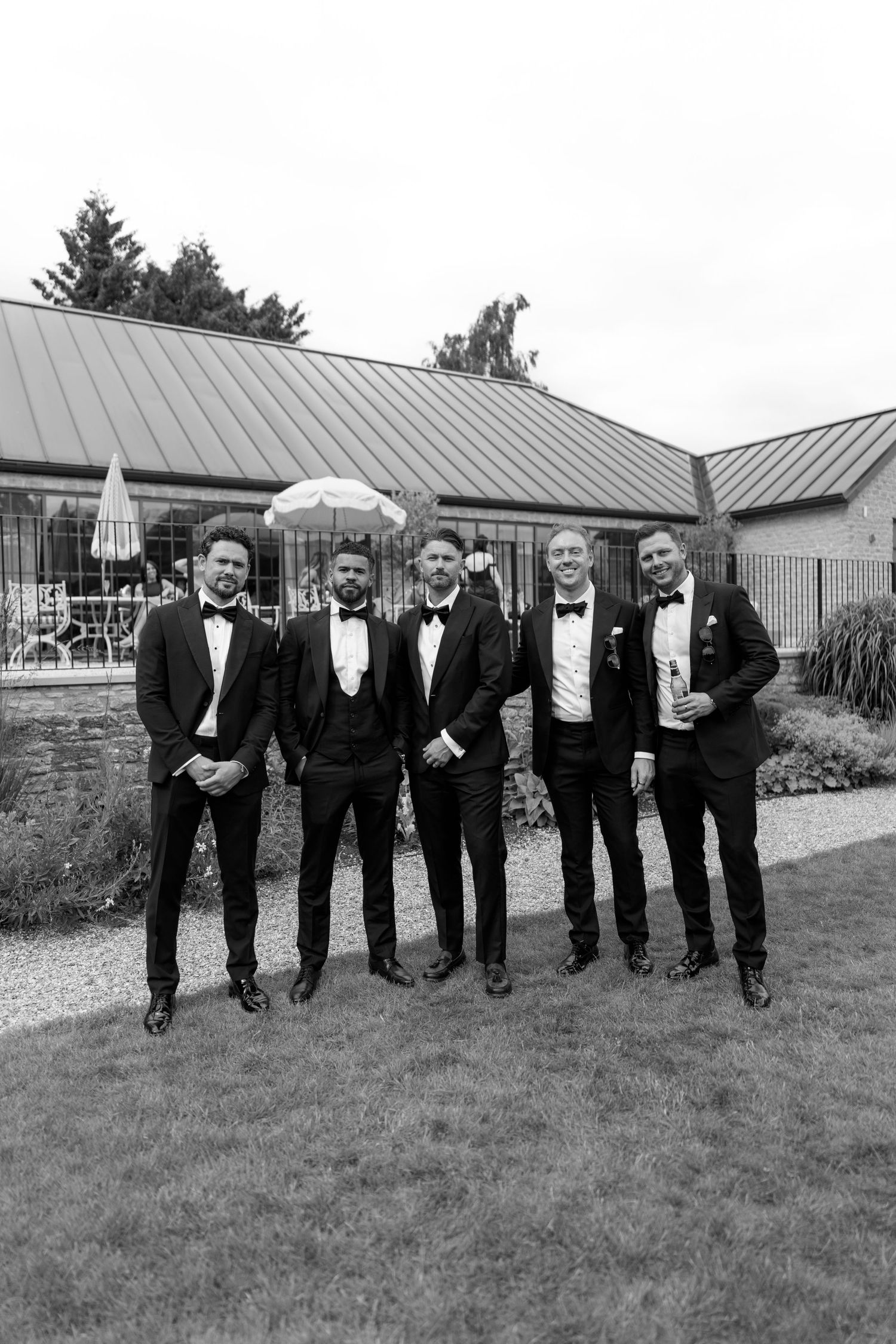 Five groomsmen in black tuxedos pose together outdoors in black and white photo.