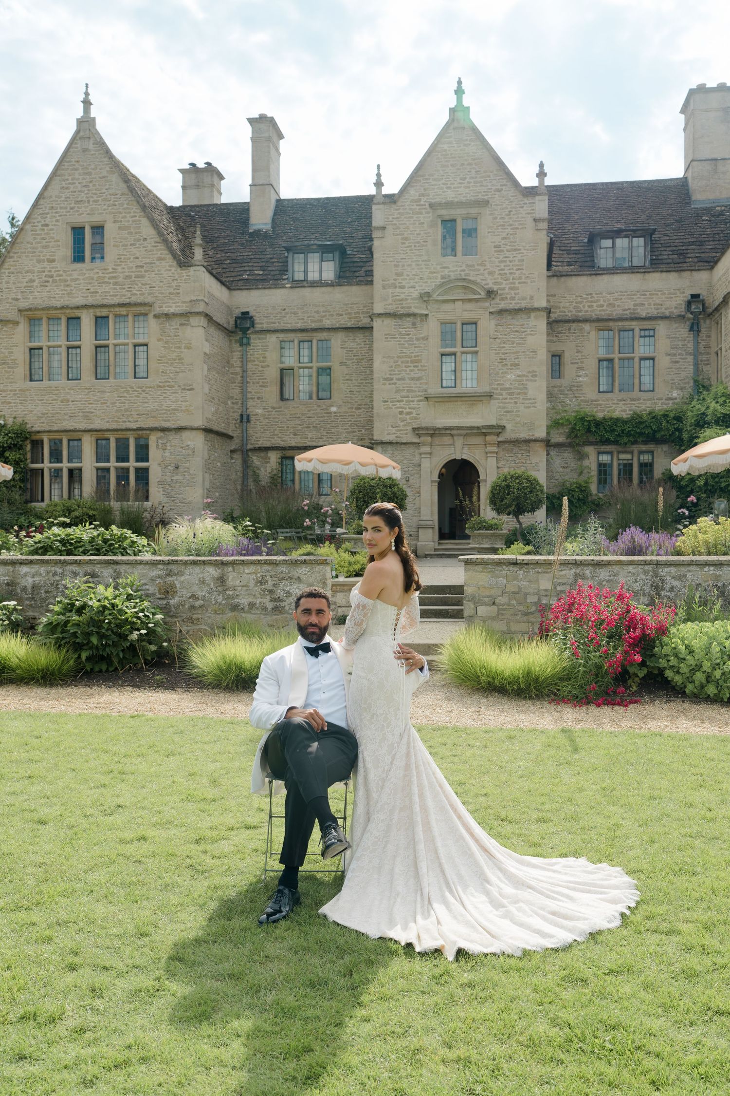 Bride holding parasol stands beside seated groom on lawn before a grand English country manor.