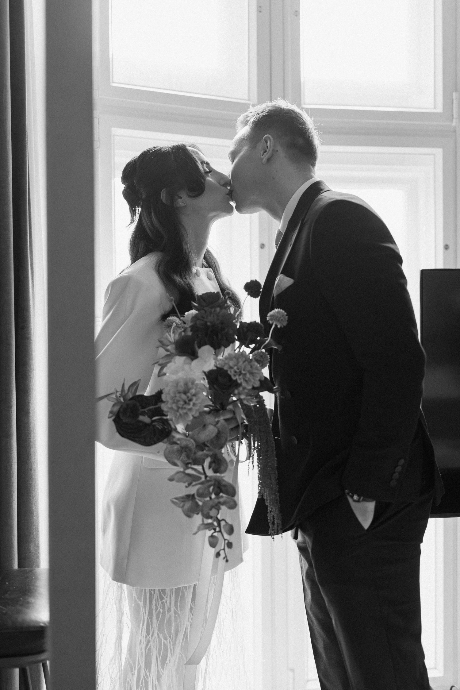 Black and white photo of bride and groom sharing a kiss while holding wedding bouquet by window.