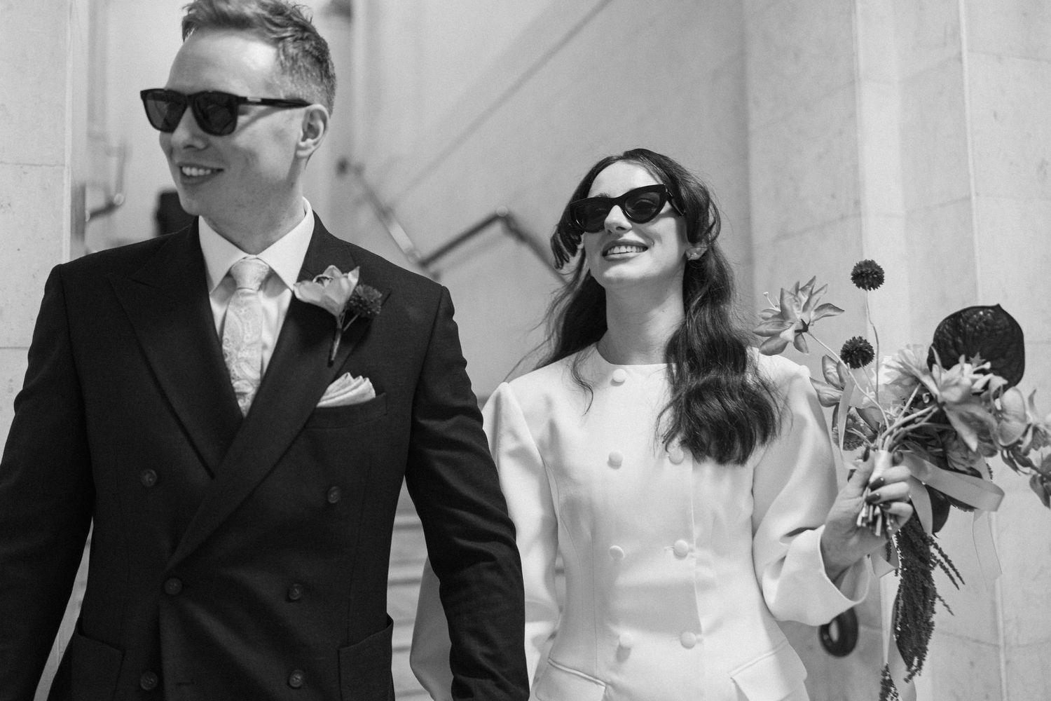 Black and white photo of stylish bride and groom walking in sunglasses, bride holding bouquet on stone steps.