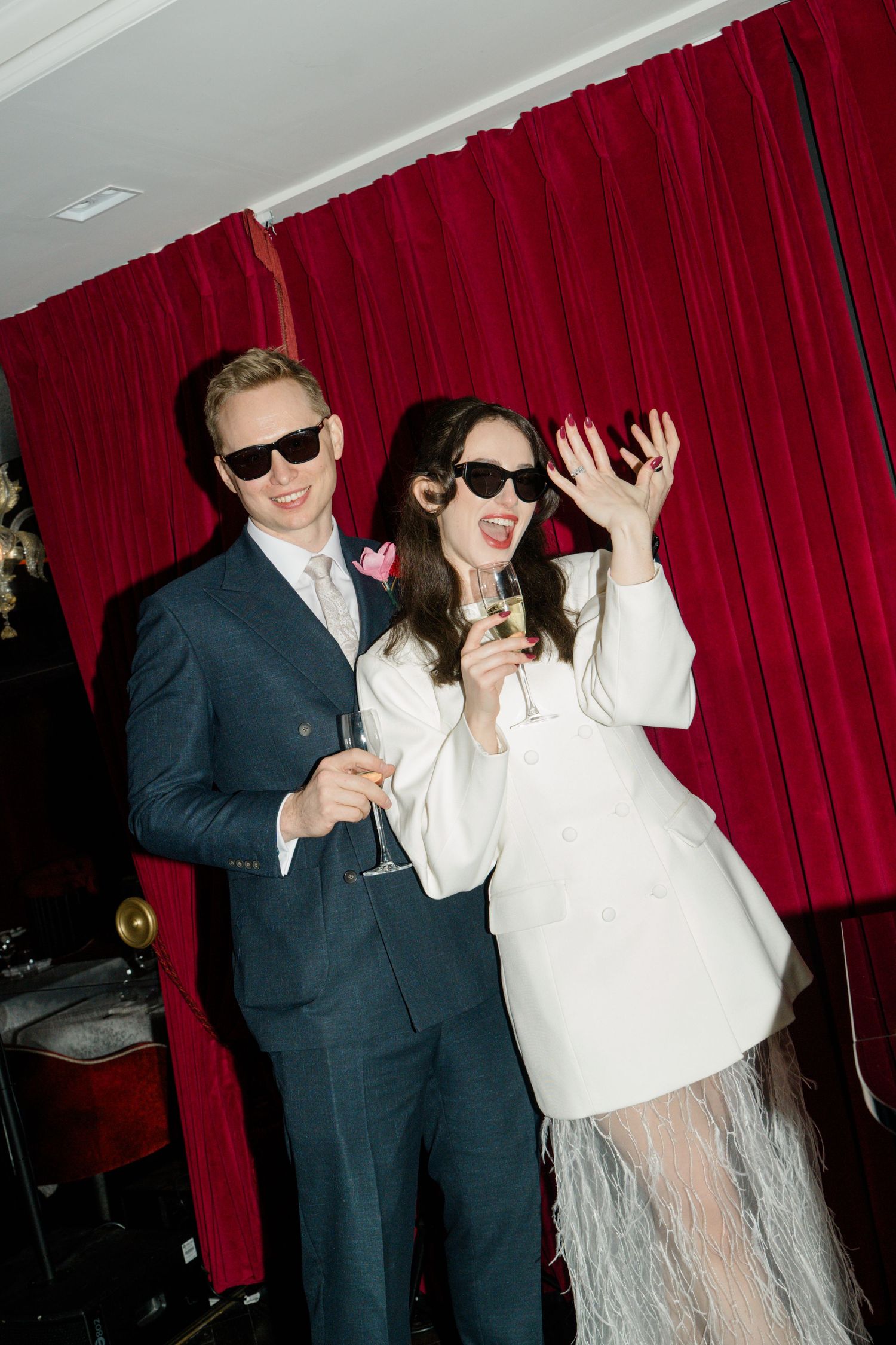 Joyful newlyweds in sunglasses celebrate with champagne glasses before dramatic red velvet curtains.