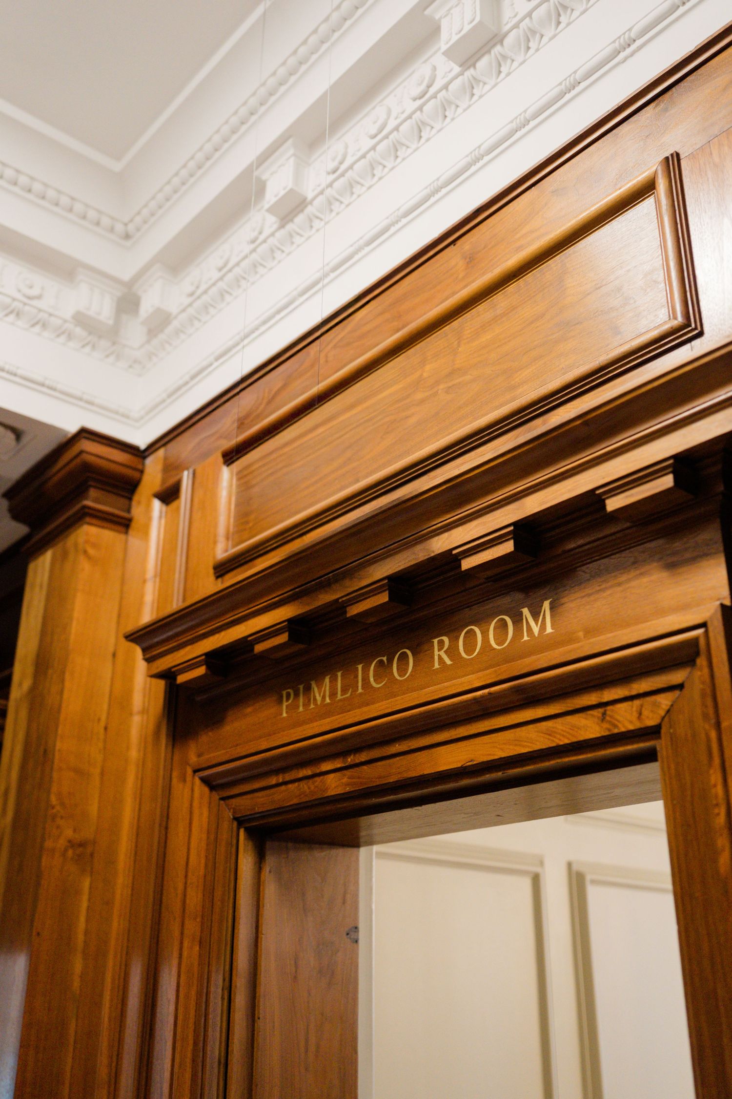Elegant wooden doorframe with gold lettering reading Pimlico Room beneath ornate white ceiling.