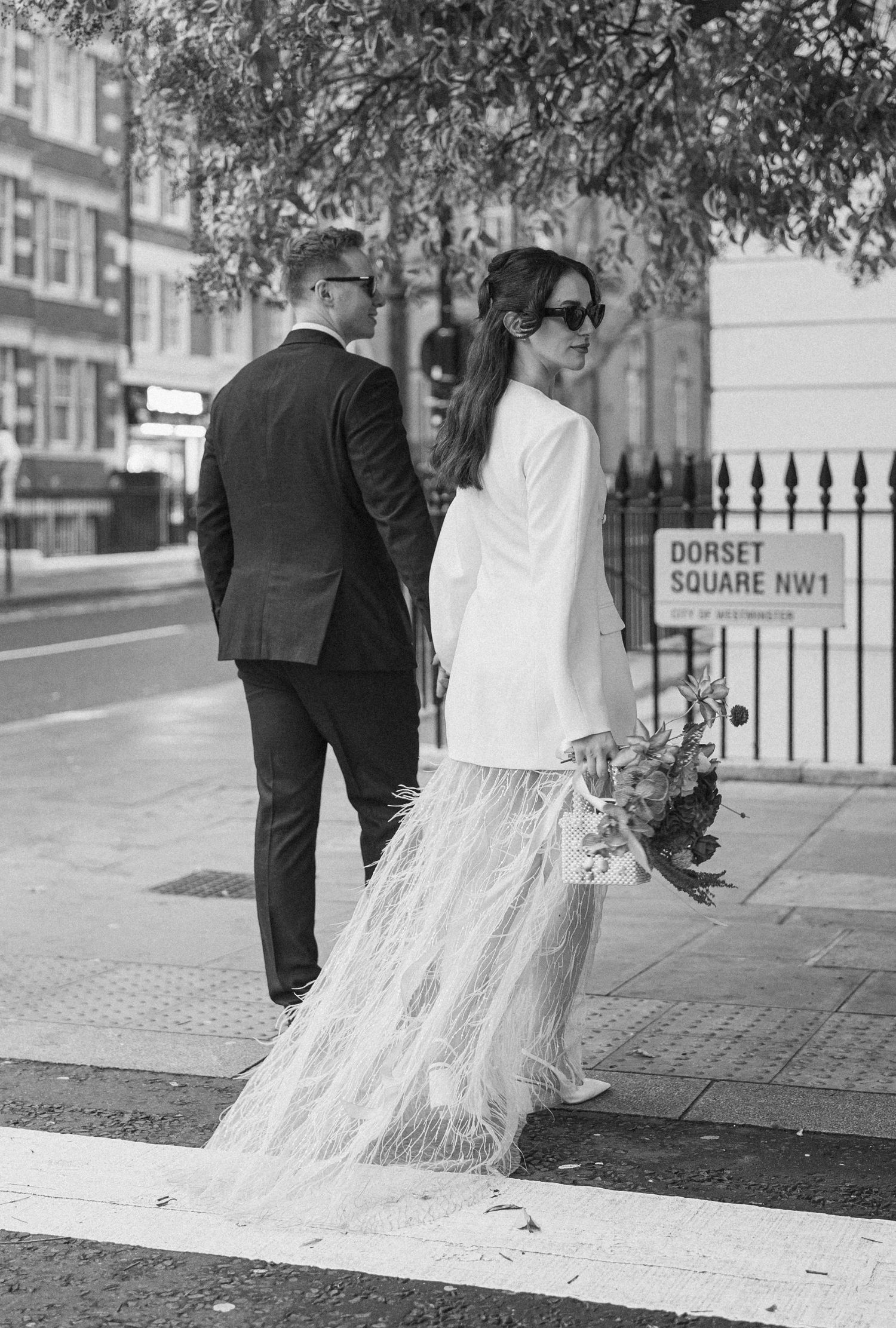 Black and white photo of bride in feather skirt crossing street near Dorset Square NW1 sign in London.