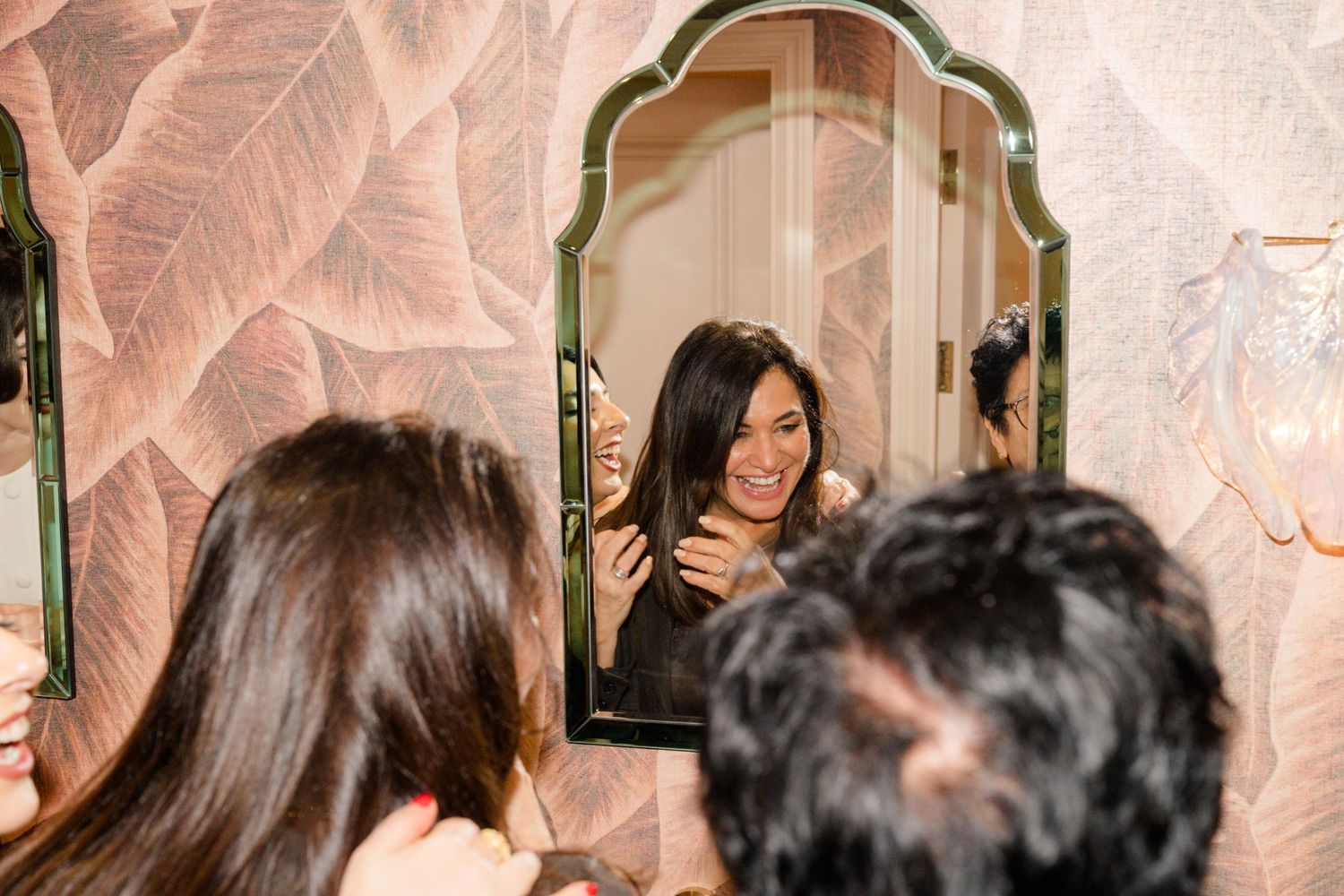 Smiling bride admires her reflection in ornate mirror with floral wallpaper background.