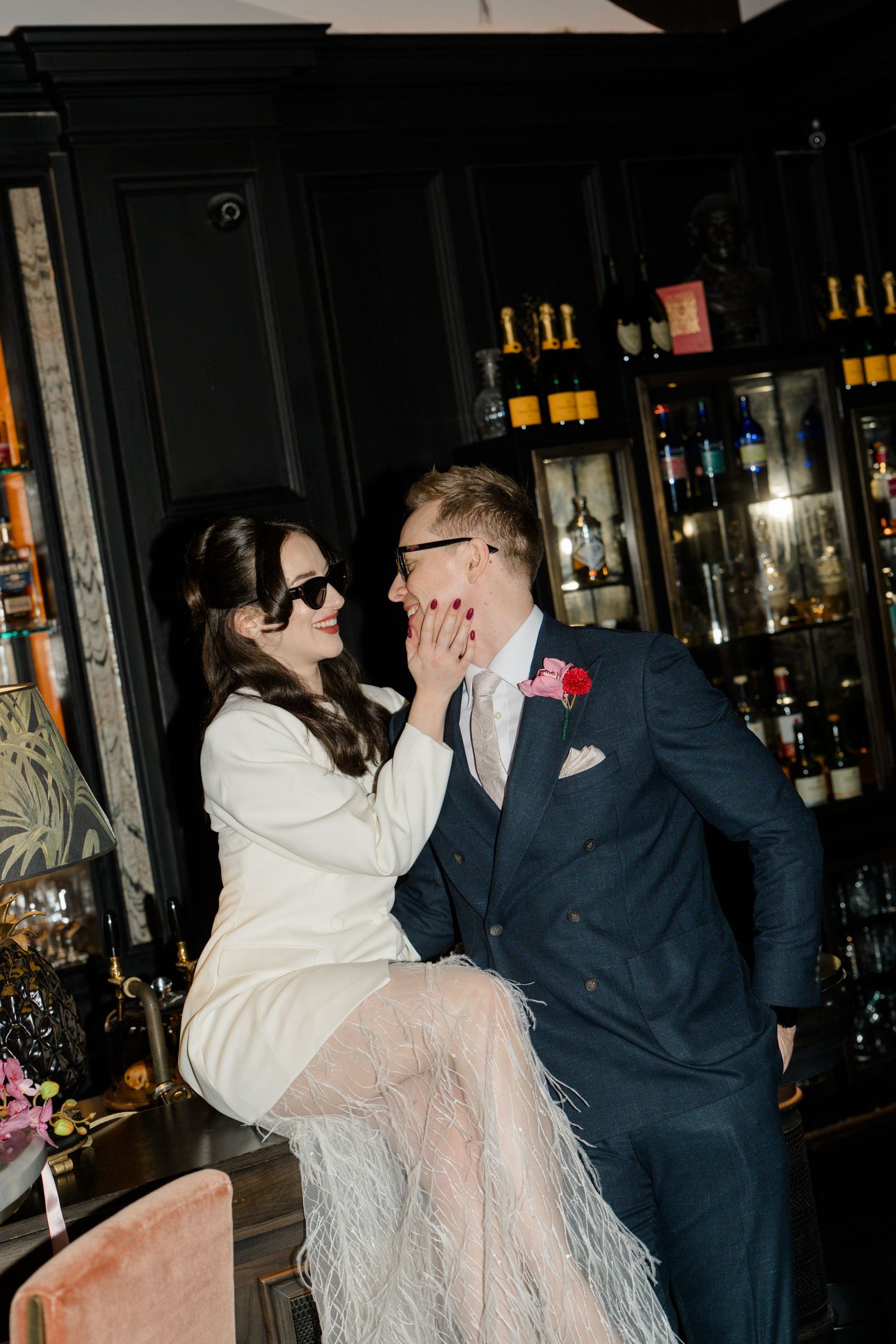 Bride and groom share a playful moment at a bar, both wearing sunglasses on their wedding day.