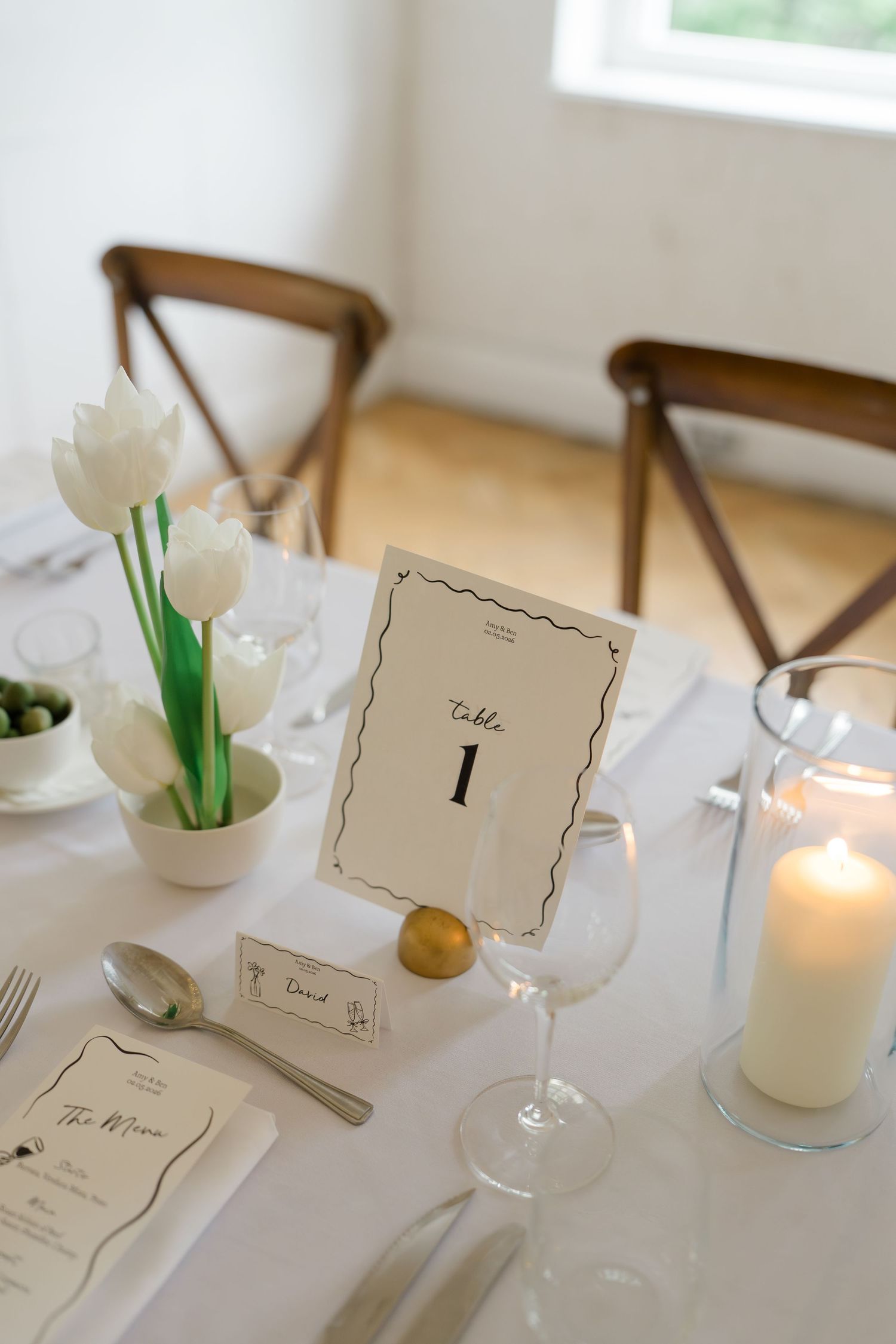 Elegant wedding table setting with white tulips, Table 1 card, candle, and place settings on white linen.
