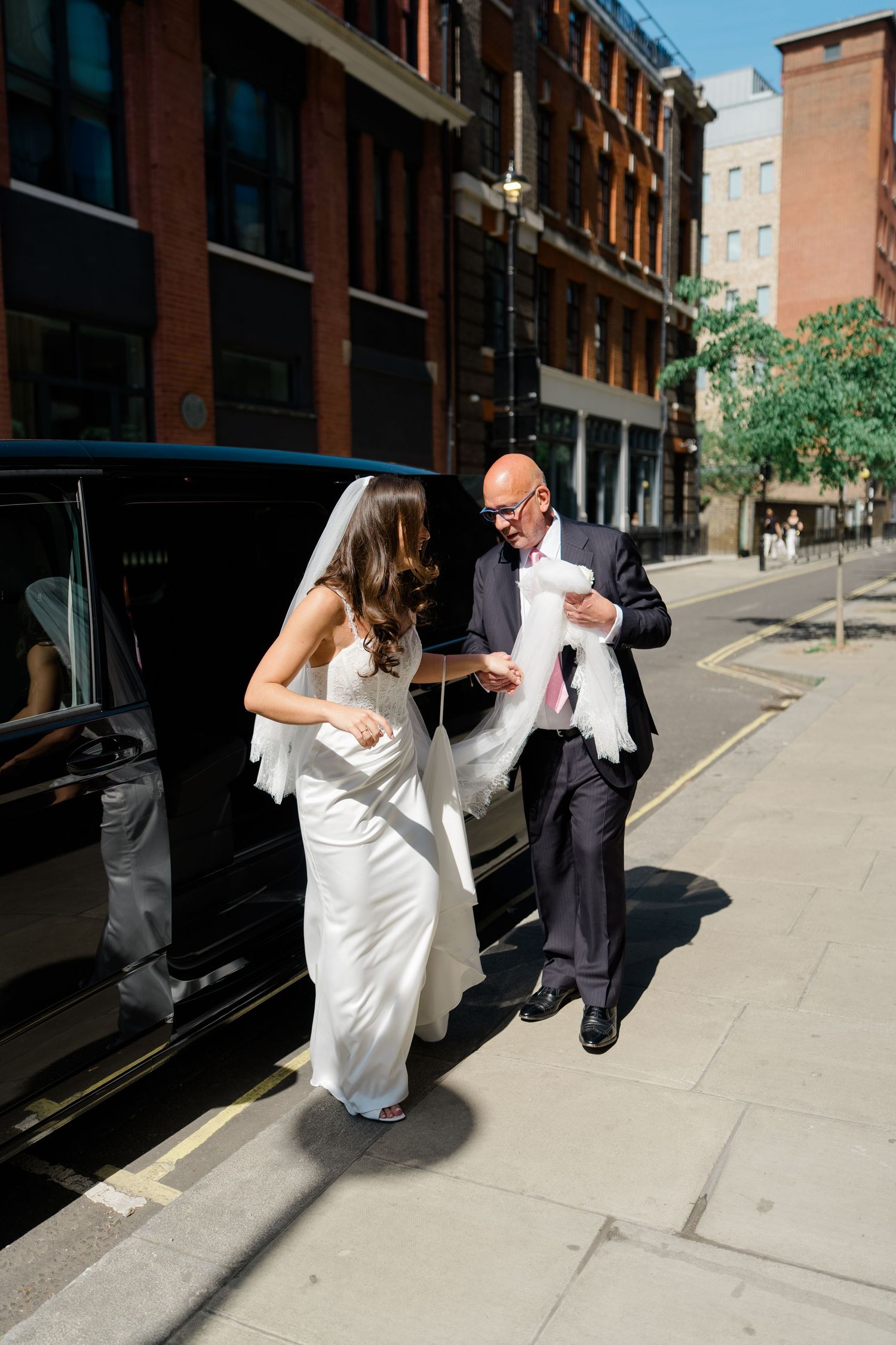 Bride in white gown and veil exits black car assisted by bald man in dark suit on sunny city street.