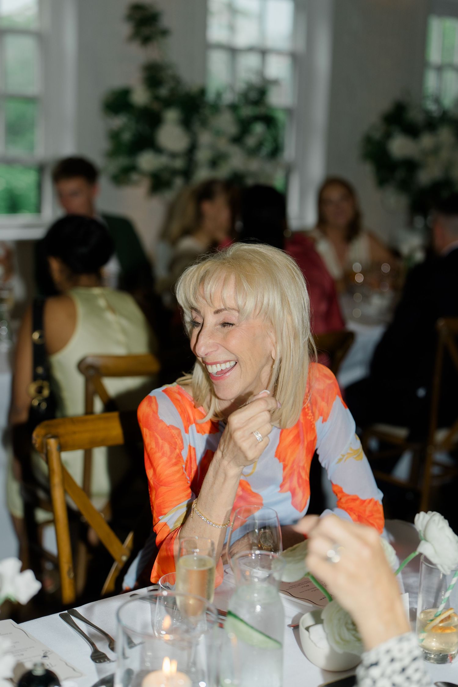 Smiling blonde woman in colorful floral dress enjoying a wedding reception dinner with guests in background.