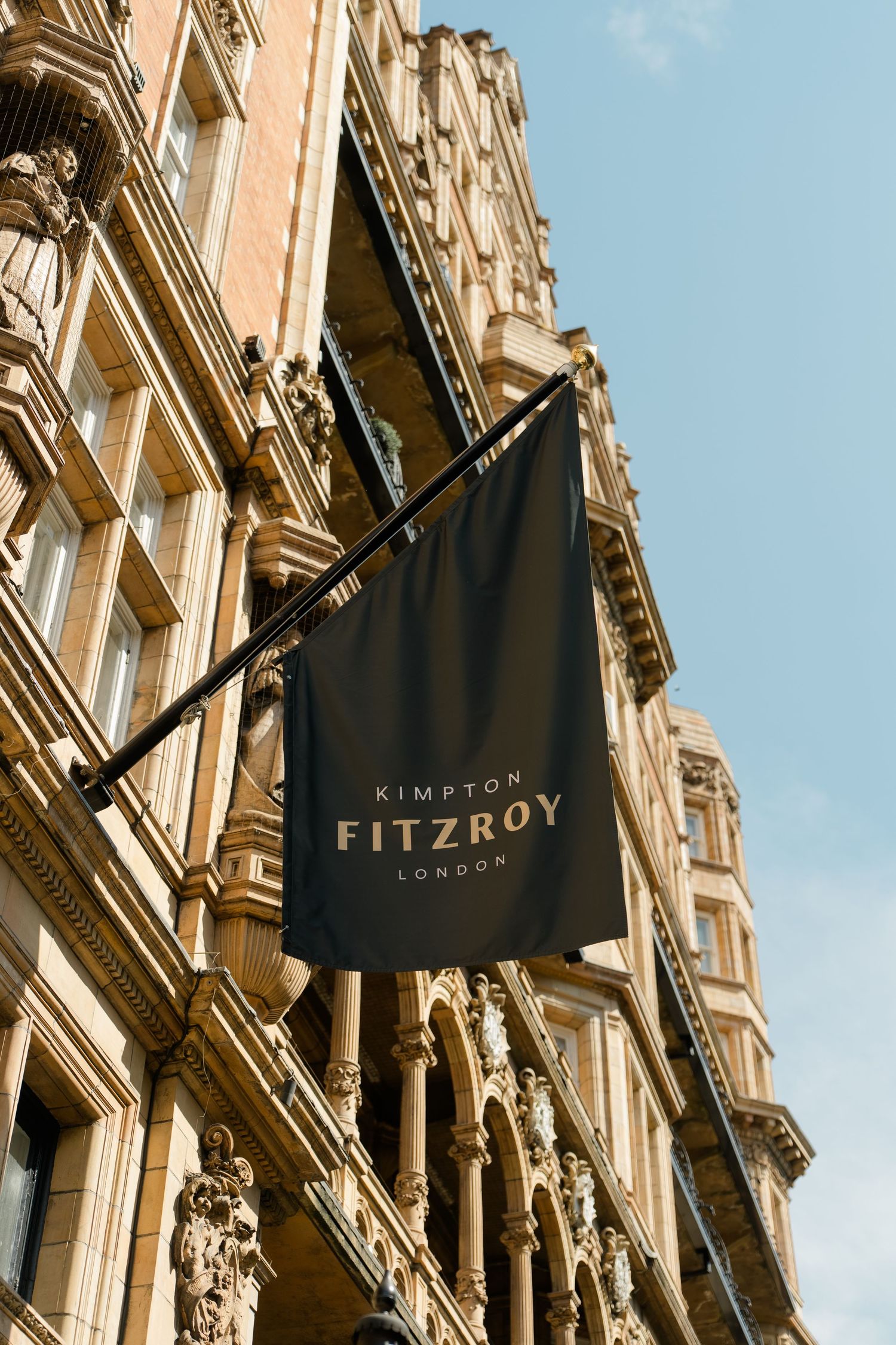 Black flag sign for Kimpton Fitzroy London hotel on ornate Victorian stone building facade against blue sky.