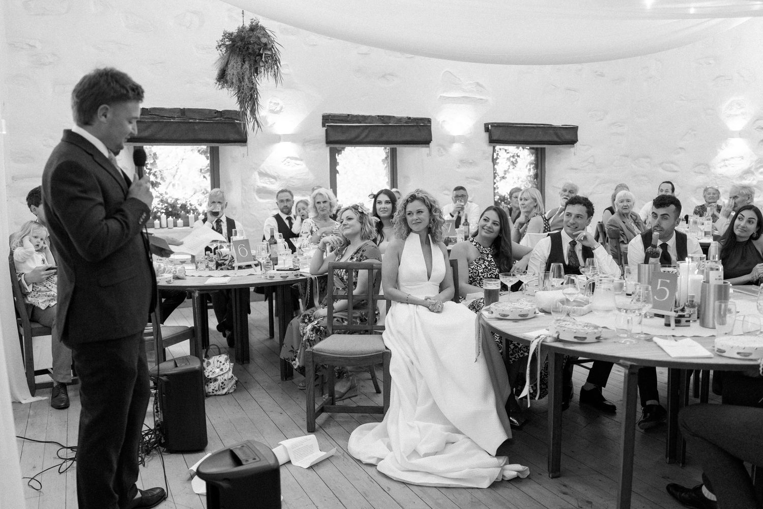 Black and white photo of wedding reception speech with guests seated at decorated tables.
