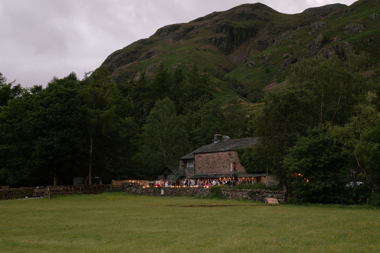 Stone building nestled at the foot of a mountain with wedding reception gathering outside.