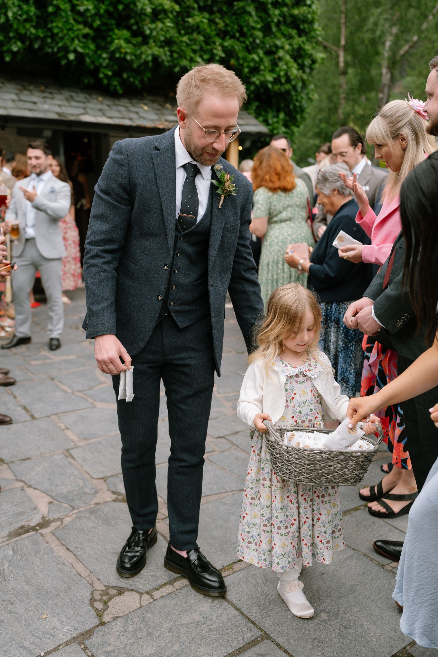 A small child in a floral dress holds hands with someone in formal attire during an outdoor event.
