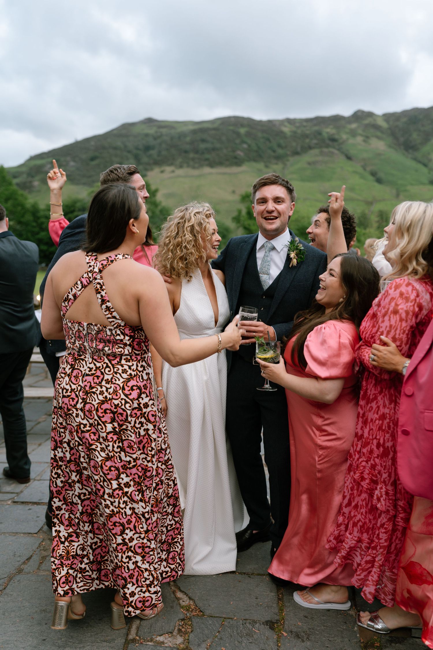 Wedding party guests in coral and floral dresses dancing together outdoors with mountains visible.