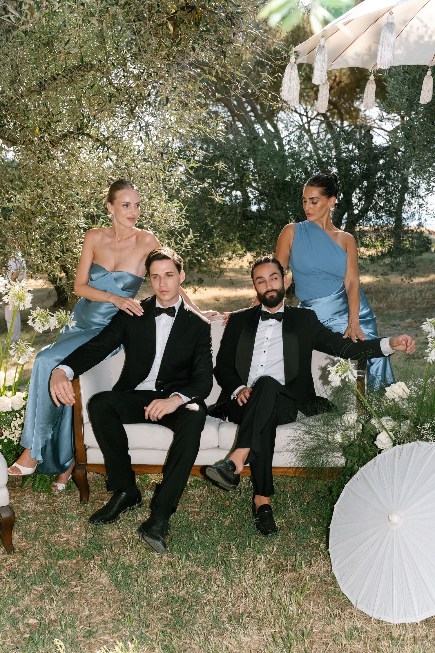 Wedding party lounges playfully on grassy hillside in coordinated blue dresses and black suits.