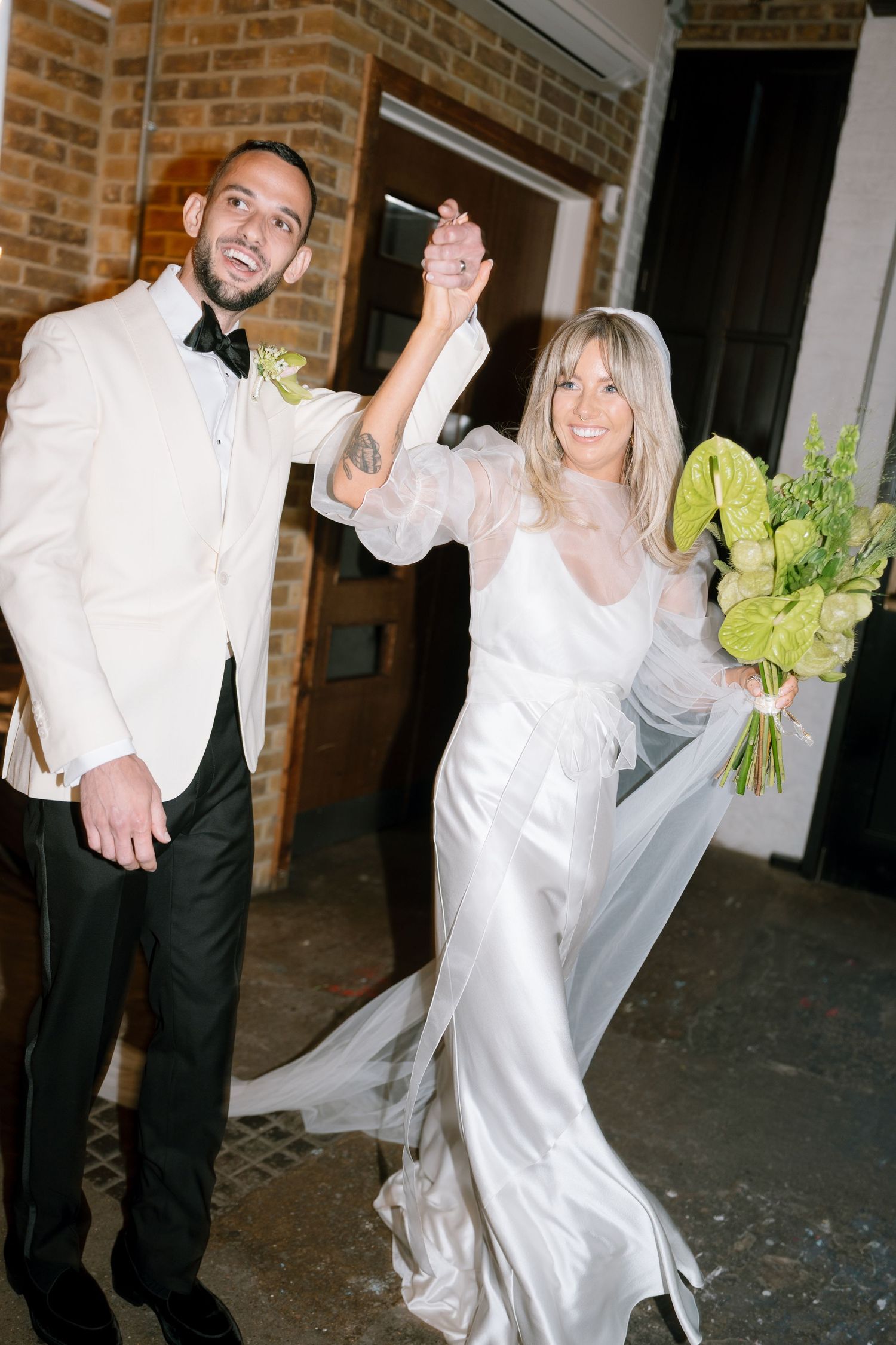 Couple dancing and twirling in an elegant white dress and formal attire against a brick wall backdrop.