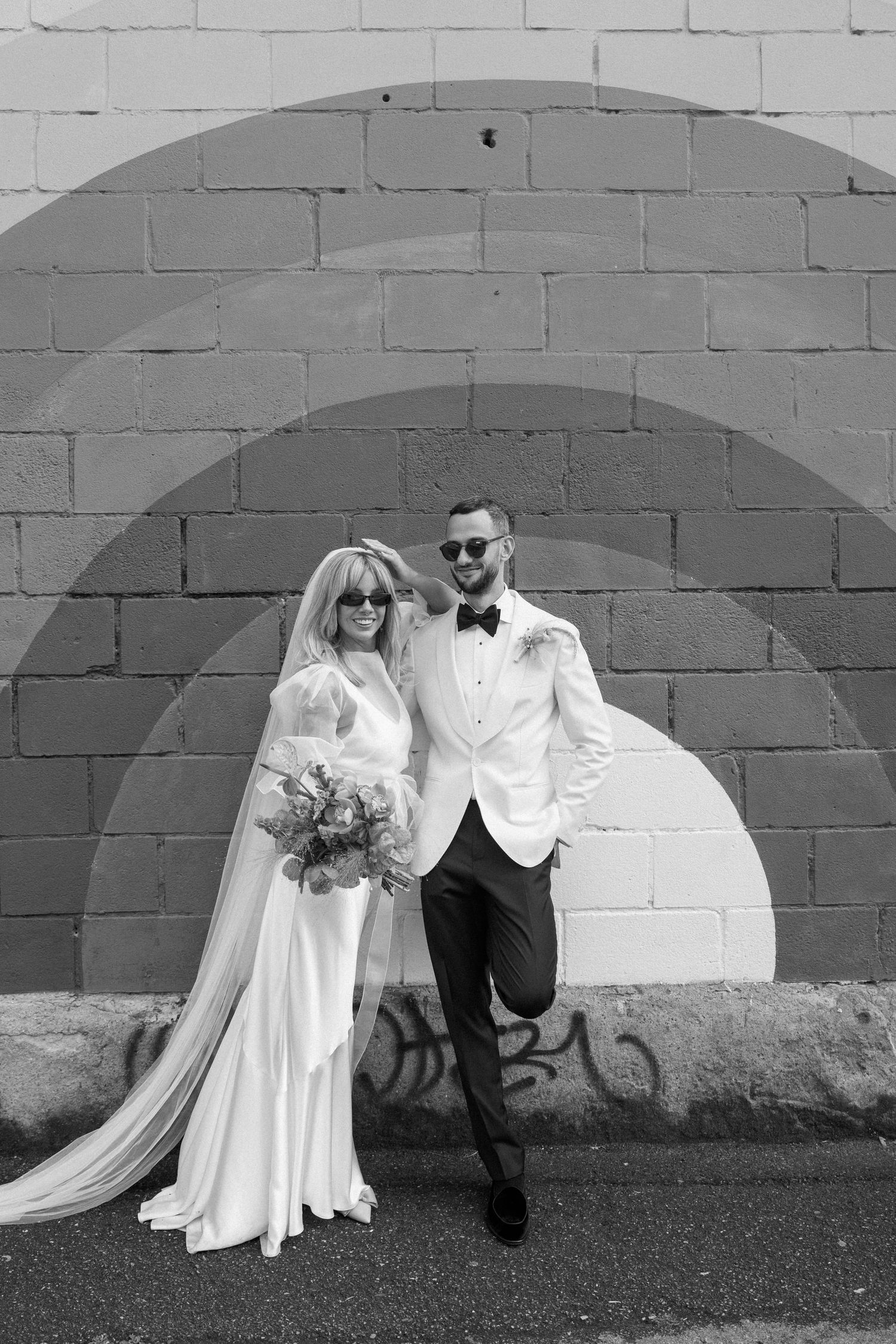 Black and white wedding portrait against white brick wall with arched detail.
