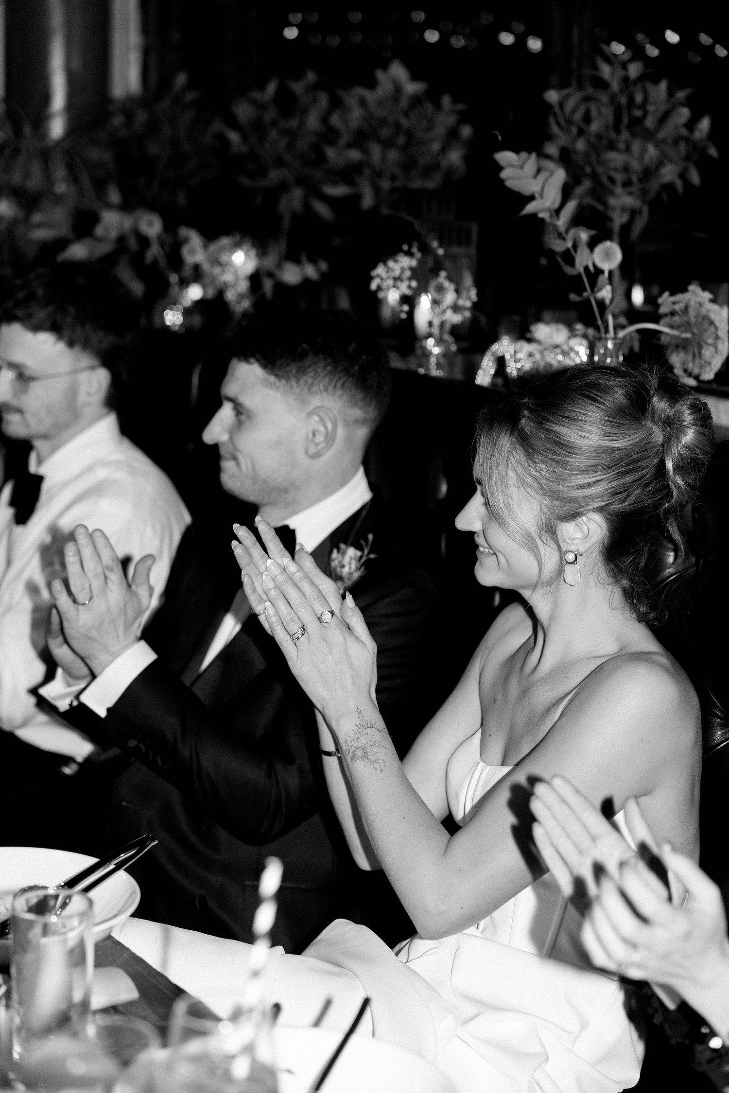 Wedding guests and a bride in formal attire applaud during a reception celebration in black and white photography.