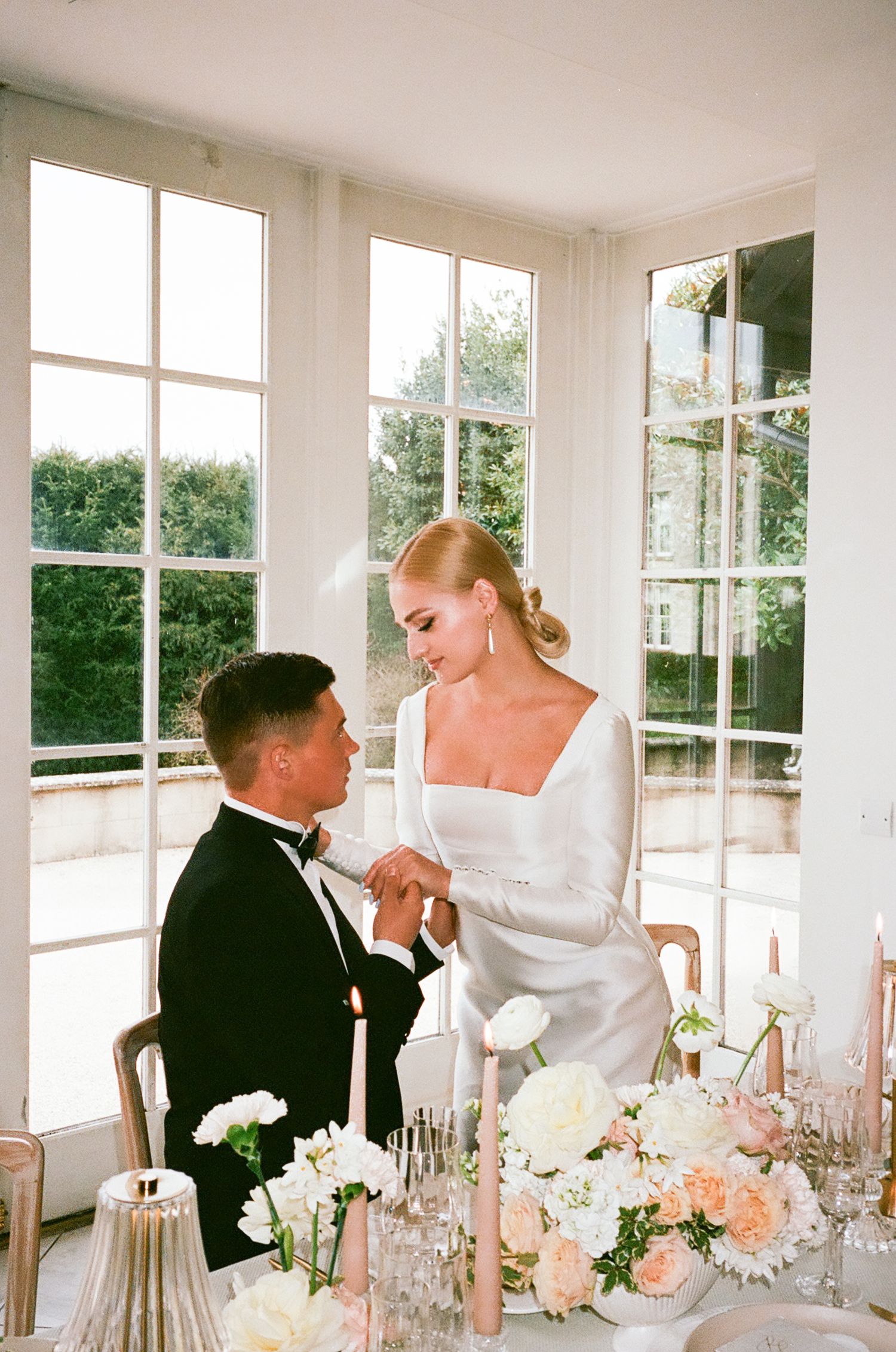 Elegant wedding reception table setting with white and peach flowers in a bright sunlit room with large windows.