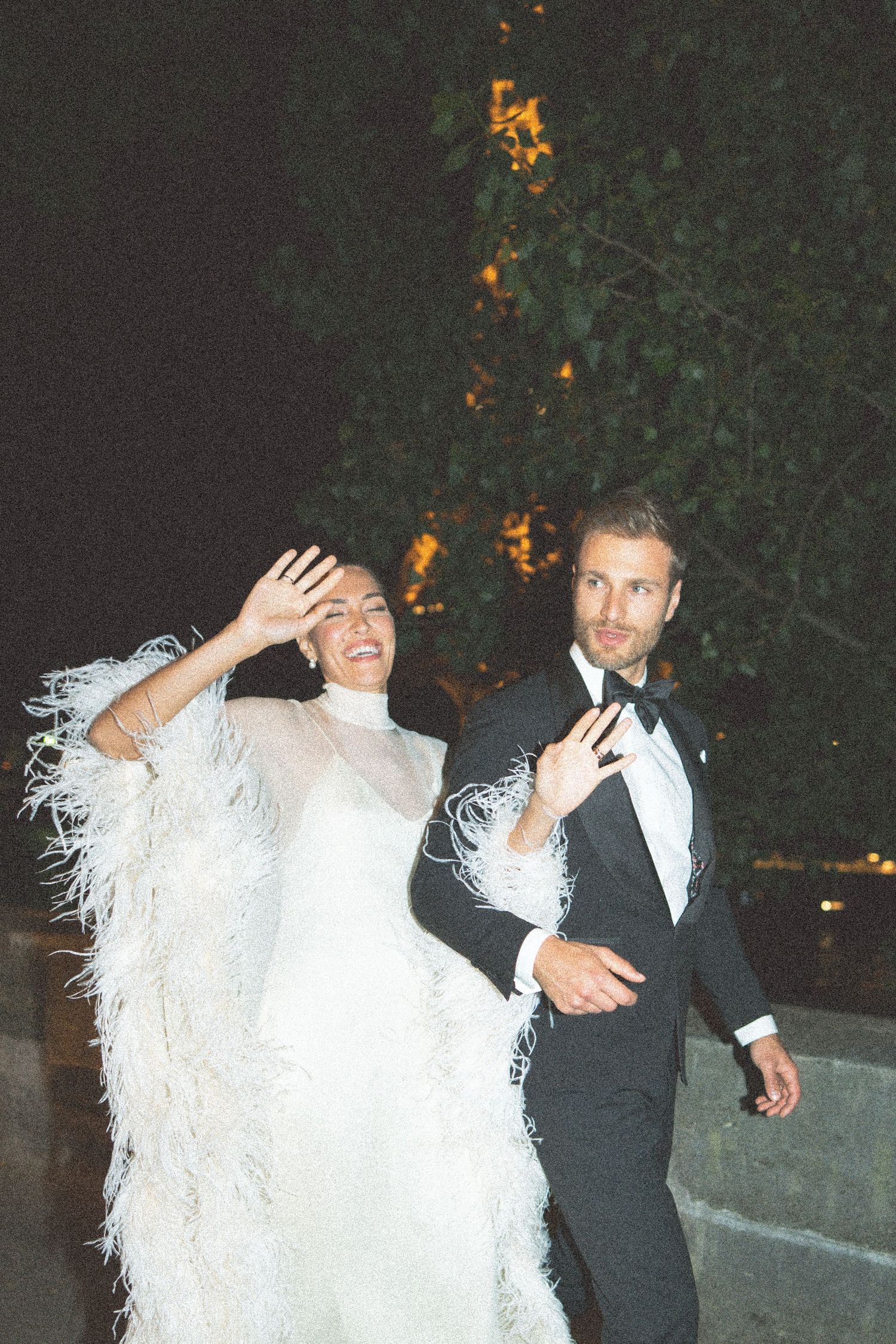 A couple in formal attire, with the bride wearing a white feathered dress and groom in black tuxedo, dancing at their wedding.