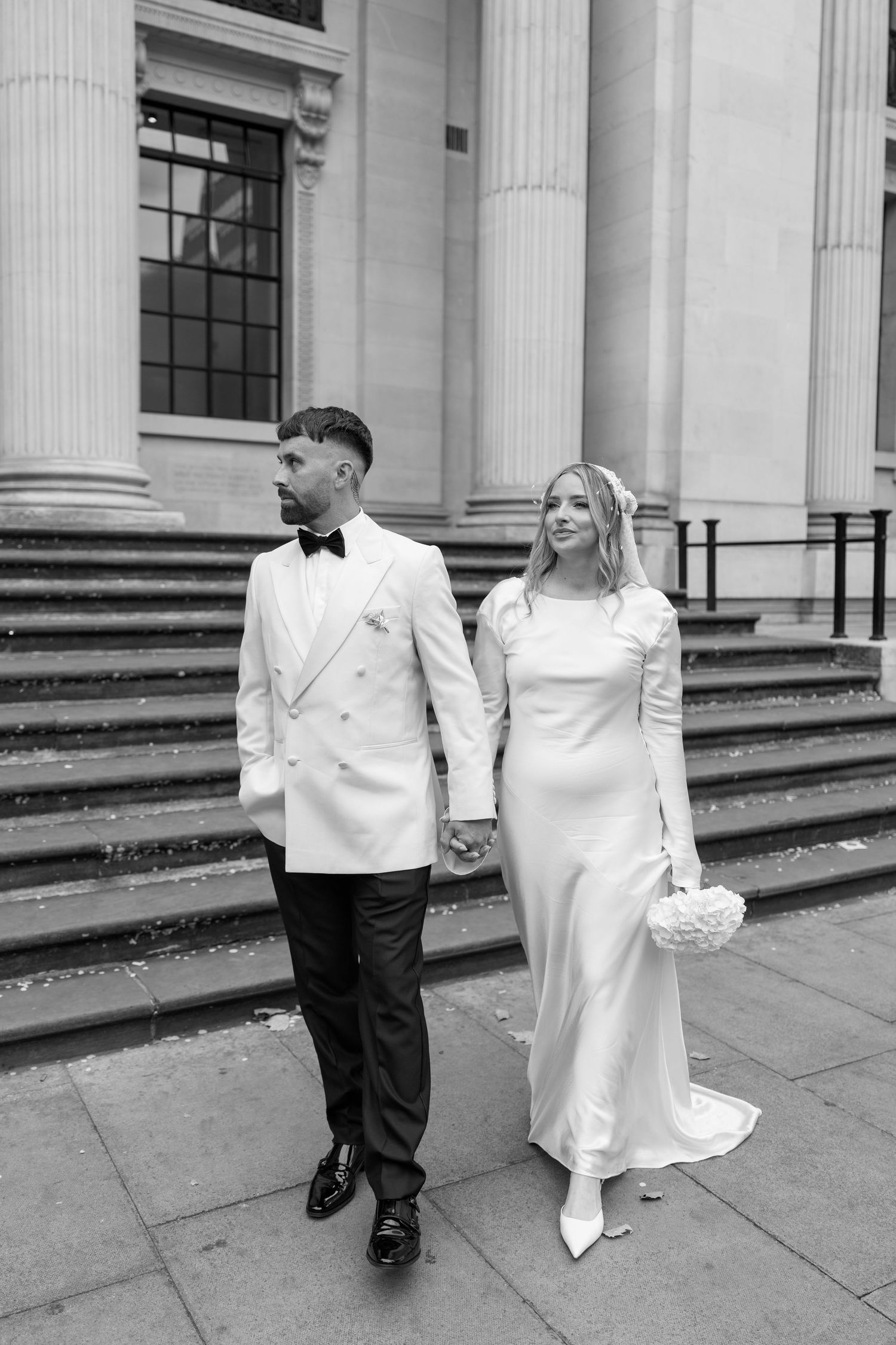 A couple in elegant white wedding attire poses on stone steps of a neoclassical building.