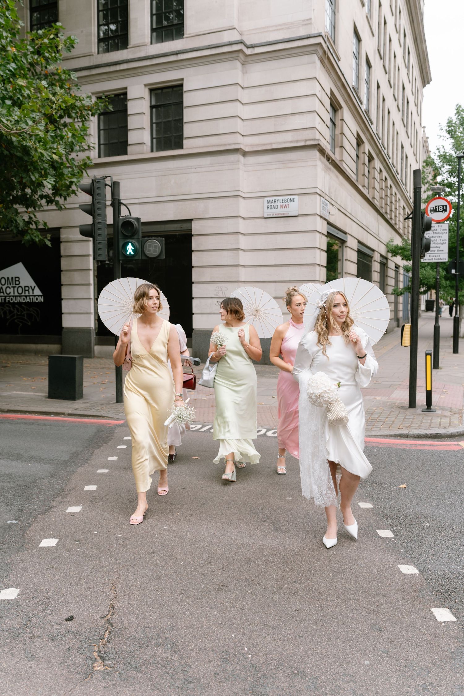 Four people in light colored dresses walk down a city street holding white parasols on a sunny day.