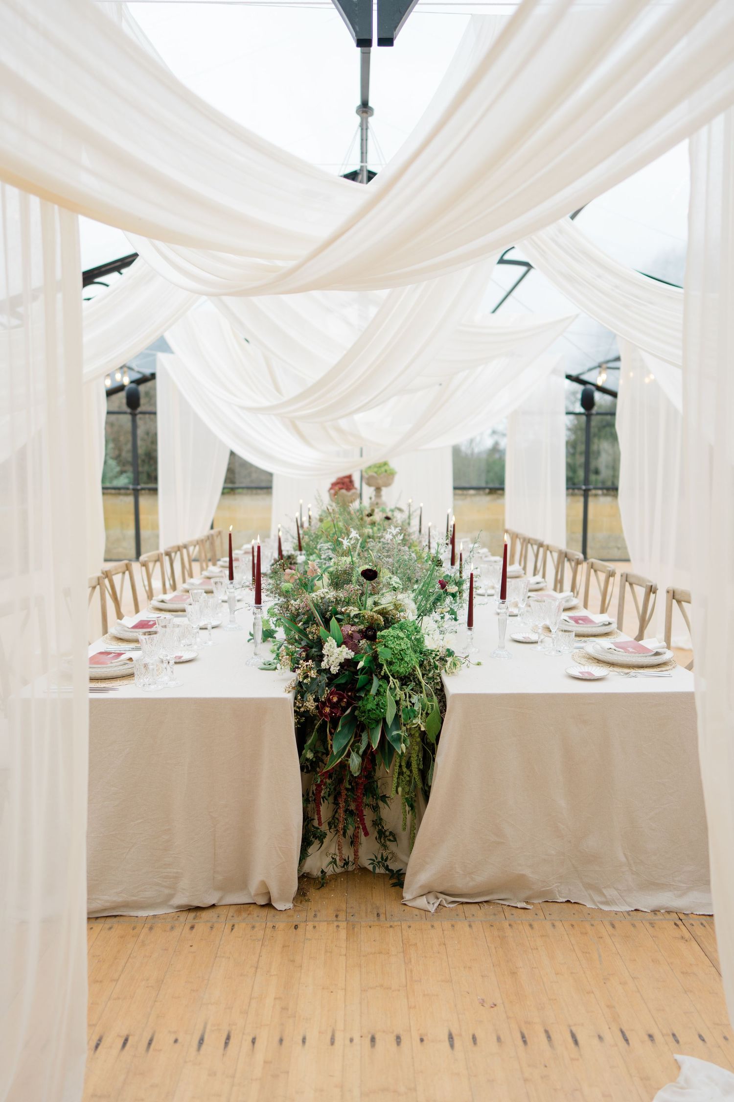 Elegant white draped tent interior with long dining tables set for a wedding reception featuring floral centerpieces.