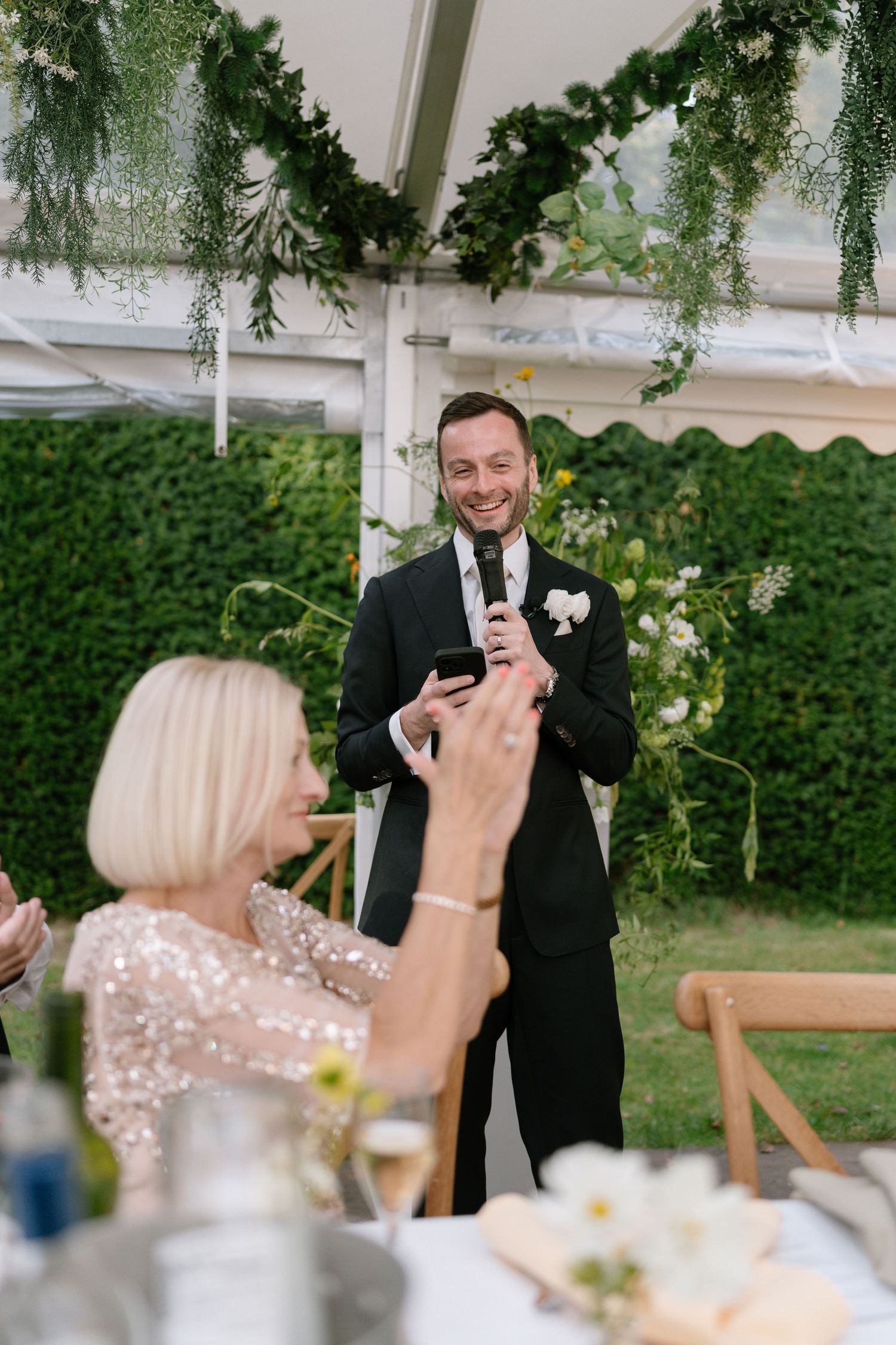 Speech being given at an outdoor wedding reception under a tent with greenery decorations.
