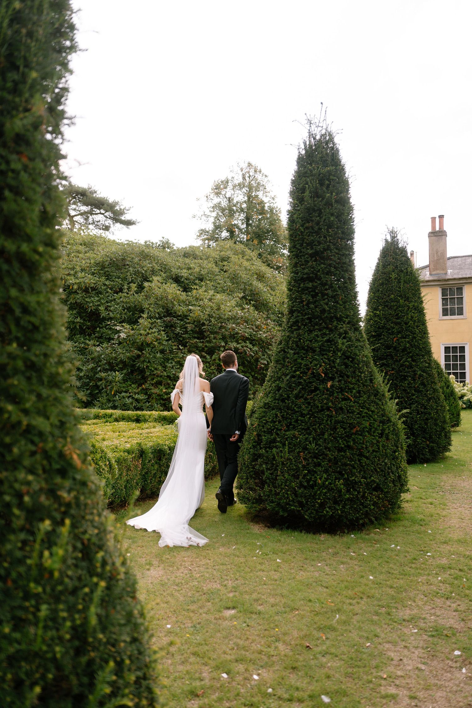 Wedding couple walks through a landscaped garden with tall cypress trees and a yellow manor house.